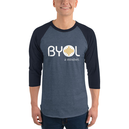 A young man wearing a heather blue and navy blue "BYOL: a mindset" raglan T-shirt with three-quarter sleeves, smiling and standing with his arms at his sides. The T-shirt features the "BYOL" logo in white and yellow on the front and is available in multiple colors (red, black, white, green, plus more) and sizes (S-3XL). The "be you out loud" logo is displayed at the top.