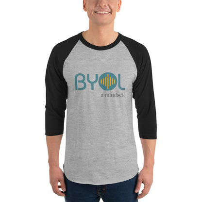 A young man wearing a heather grey and black "BYOL: a mindset" raglan T-shirt with three-quarter sleeves, smiling and standing with his arms at his sides. The T-shirt features the "BYOL" logo in teal and yellow on the front and is available in multiple colors (red, black, white, green, plus more) and sizes (S-3XL). The "be you out loud" logo is displayed at the top.