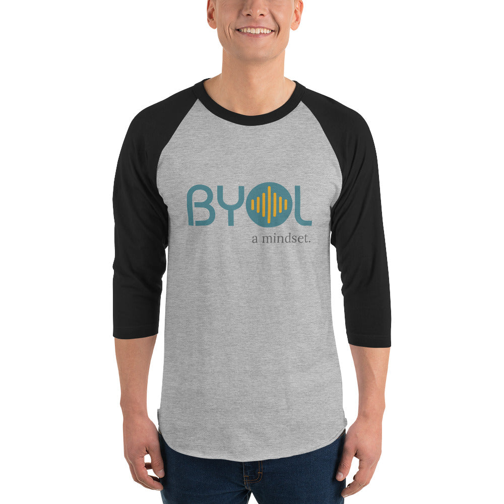 A young man wearing a gray and black "BYOL: a mindset" raglan T-shirt with three-quarter sleeves, smiling and standing with his arms at his sides. The T-shirt features the "BYOL" logo in teal and yellow on the front and is available in multiple colors (red, black, white, green, plus more) and sizes (S-3XL). The "be you out loud" logo is displayed at the top.