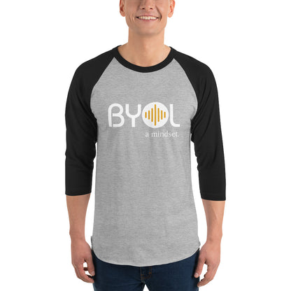 A young man wearing a gray and black "BYOL: a mindset" raglan T-shirt with three-quarter sleeves, smiling and standing with his arms at his sides. The T-shirt features the "BYOL" logo in white and yellow on the front and is available in multiple colors (red, black, white, green, plus more) and sizes (S-3XL). The "be you out loud" logo is displayed at the top.