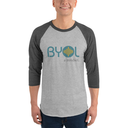 A young man wearing a heather grey and heather dark grey "BYOL: a mindset" raglan T-shirt with three-quarter sleeves, smiling and standing with his arms at his sides. The T-shirt features the "BYOL" logo in teal and yellow on the front and is available in multiple colors (red, black, white, green, plus more) and sizes (S-3XL). The "be you out loud" logo is displayed at the top.