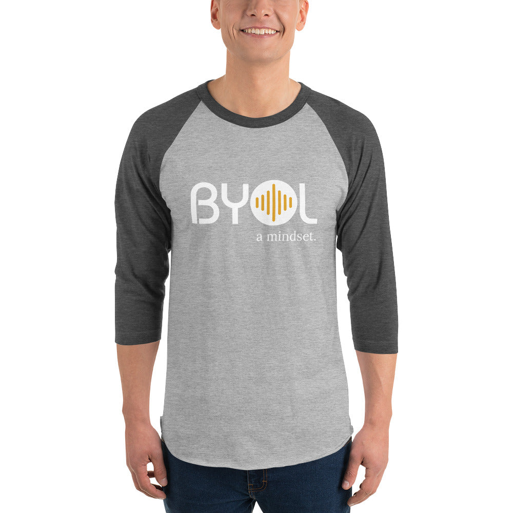 A young man wearing a gray and dark grey "BYOL: a mindset" raglan T-shirt with three-quarter sleeves, smiling and standing with his arms at his sides. The T-shirt features the "BYOL" logo in white and yellow on the front and is available in multiple colors (red, black, white, green, plus more) and sizes (S-3XL). The "be you out loud" logo is displayed at the top.