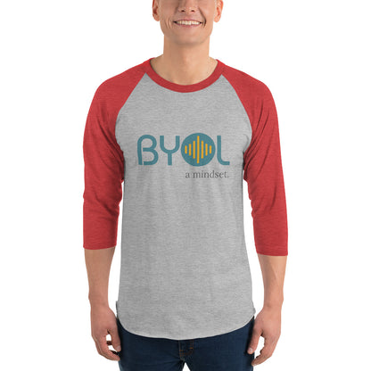 A young man wearing a heather grey and red "BYOL: a mindset" raglan T-shirt with three-quarter sleeves, smiling and standing with his arms at his sides. The T-shirt features the "BYOL" logo in teal and yellow on the front and is available in multiple colors (red, black, white, green, plus more) and sizes (S-3XL). The "be you out loud" logo is displayed at the top.