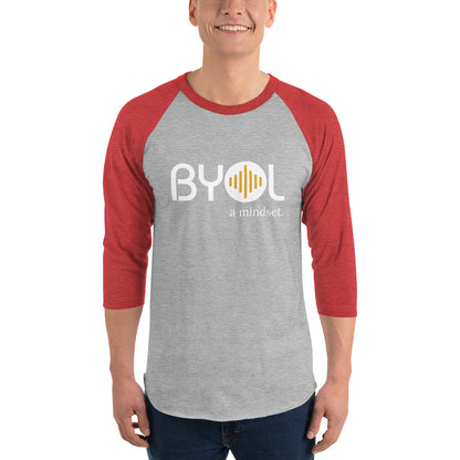 A young man wearing a gray and red "BYOL: a mindset" raglan T-shirt with three-quarter sleeves, smiling and standing with his arms at his sides. The T-shirt features the "BYOL" logo in white and yellow on the front and is available in multiple colors (red, black, white, green, plus more) and sizes (S-3XL). The "be you out loud" logo is displayed at the top.