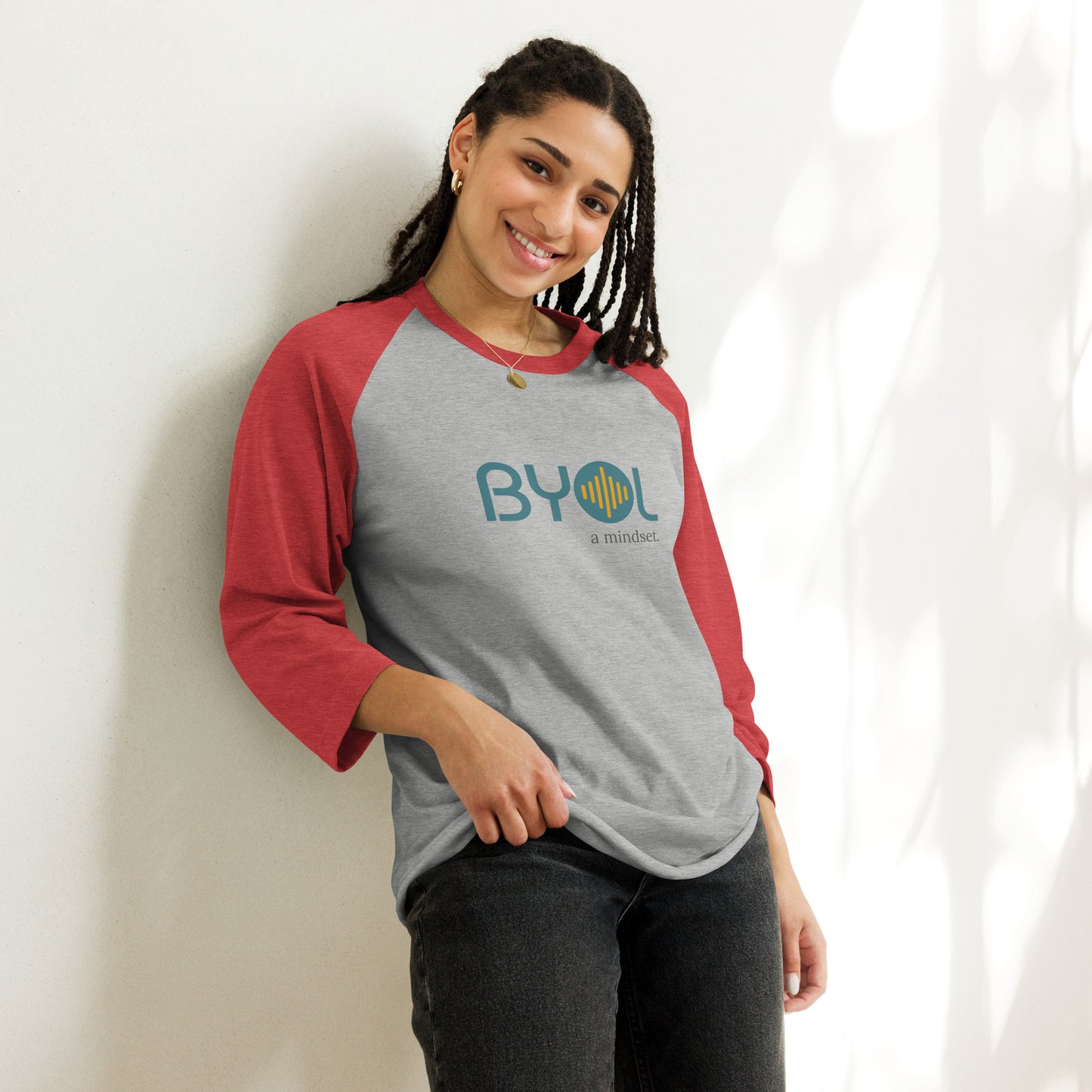 Woman wearing a heather grey and red BYOL mindset raglan t-shirt with color options in red, black, gray, and green, available in sizes S to 3XL.