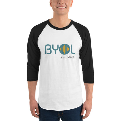 A young man wearing a white and black "BYOL: a mindset" raglan T-shirt with three-quarter sleeves, smiling and standing with his arms at his sides. The T-shirt features the "BYOL" logo in teal and yellow on the front and is available in multiple colors (red, black, white, green, plus more) and sizes (S-3XL). The "be you out loud" logo is displayed at the top.