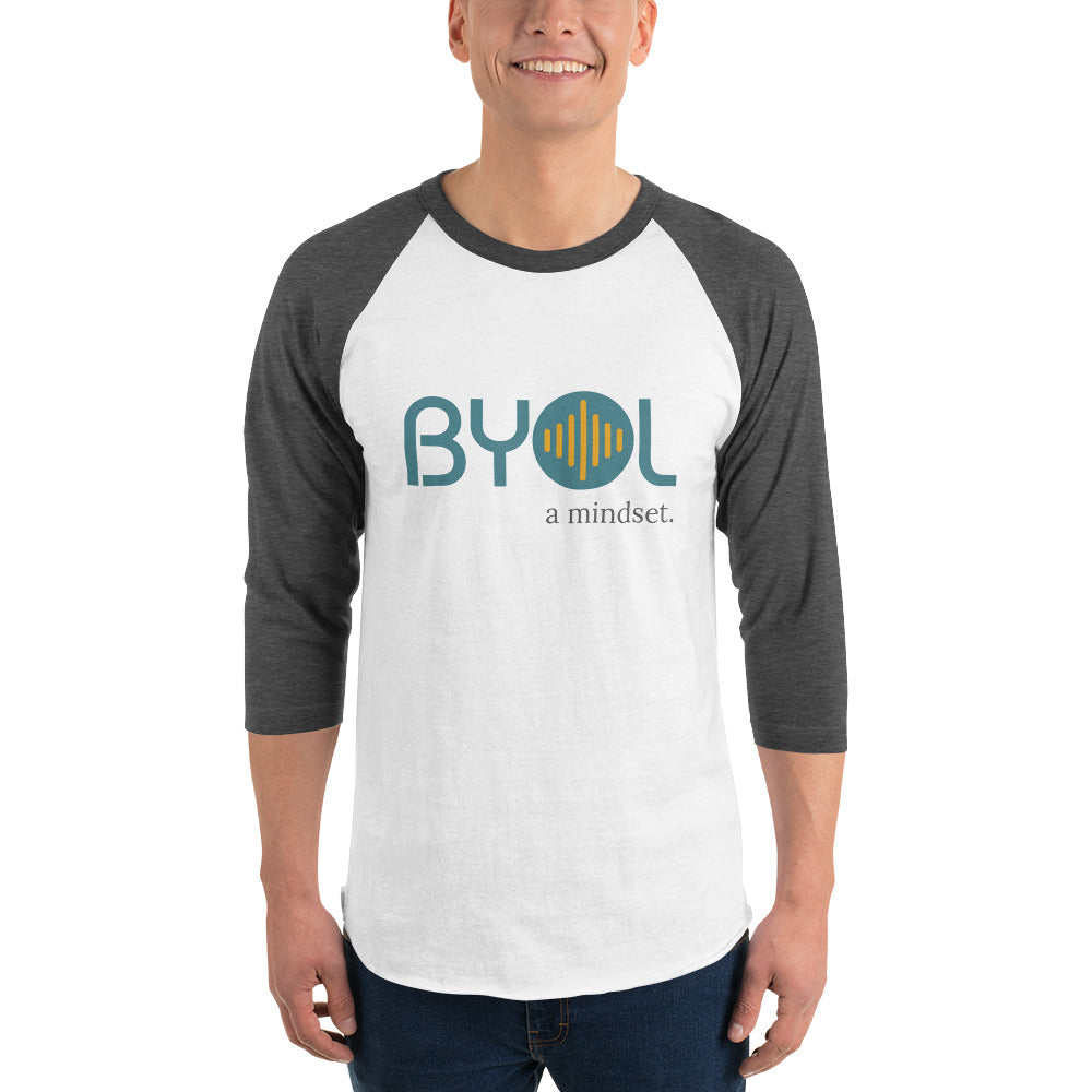 A young man wearing a white and heather dark grey "BYOL: a mindset" raglan T-shirt with three-quarter sleeves, smiling and standing with his arms at his sides. The T-shirt features the "BYOL" logo in teal and yellow on the front and is available in multiple colors (red, black, white, green, plus more) and sizes (S-3XL). The "be you out loud" logo is displayed at the top.