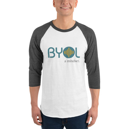 A young man wearing a white and grey "BYOL: a mindset" raglan T-shirt with three-quarter sleeves, smiling and standing with his arms at his sides. The T-shirt features the "BYOL" logo in teal and yellow on the front and is available in multiple colors (red, black, white, green, plus more) and sizes (S-3XL). The "be you out loud" logo is displayed at the top.