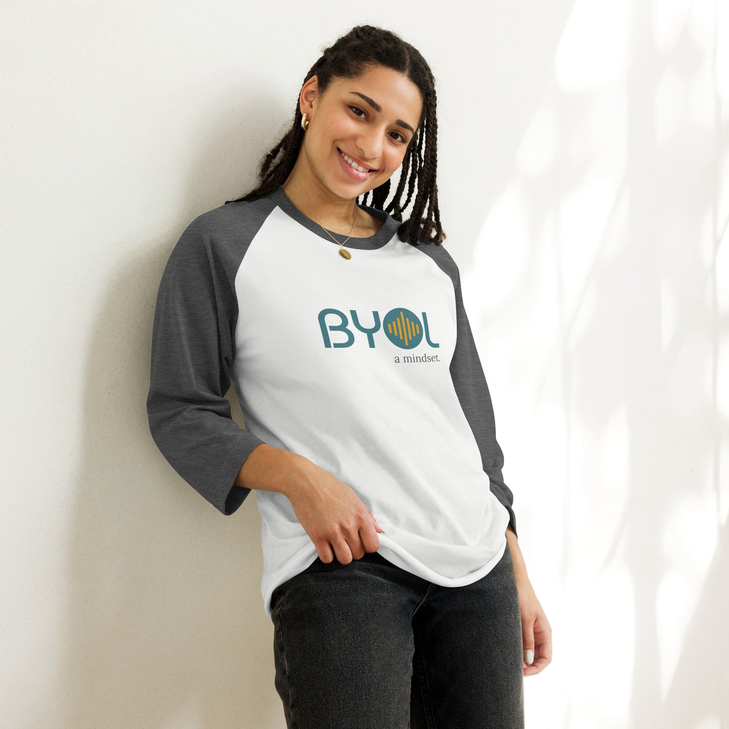 Woman wearing a white and heather dark grey BYOL mindset raglan t-shirt with color options in red, black, gray, and green, available in sizes S to 3XL.