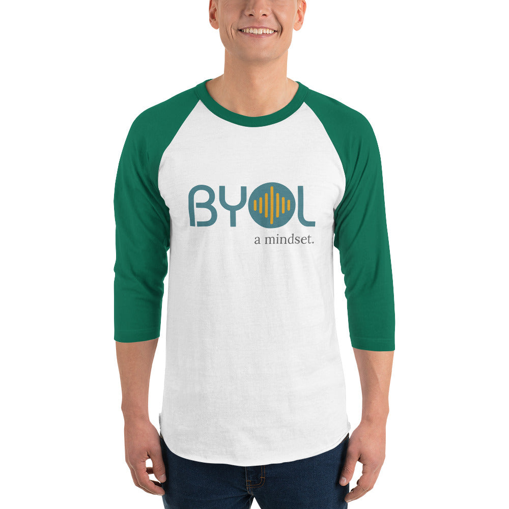 A young man wearing a white and green "BYOL: a mindset" raglan T-shirt with three-quarter sleeves, smiling and standing with his arms at his sides. The T-shirt features the "BYOL" logo in teal and yellow on the front and is available in multiple colors (red, black, white, green, plus more) and sizes (S-3XL). The "be you out loud" logo is displayed at the top.