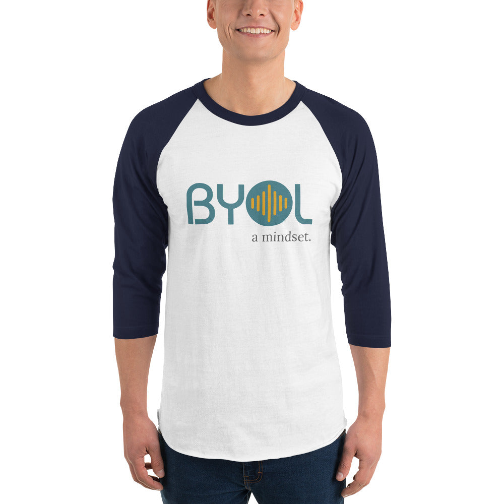 A young man wearing a white and navy blue "BYOL: a mindset" raglan T-shirt with three-quarter sleeves, smiling and standing with his arms at his sides. The T-shirt features the "BYOL" logo in teal and yellow on the front and is available in multiple colors (red, black, white, green, plus more) and sizes (S-3XL). The "be you out loud" logo is displayed at the top.