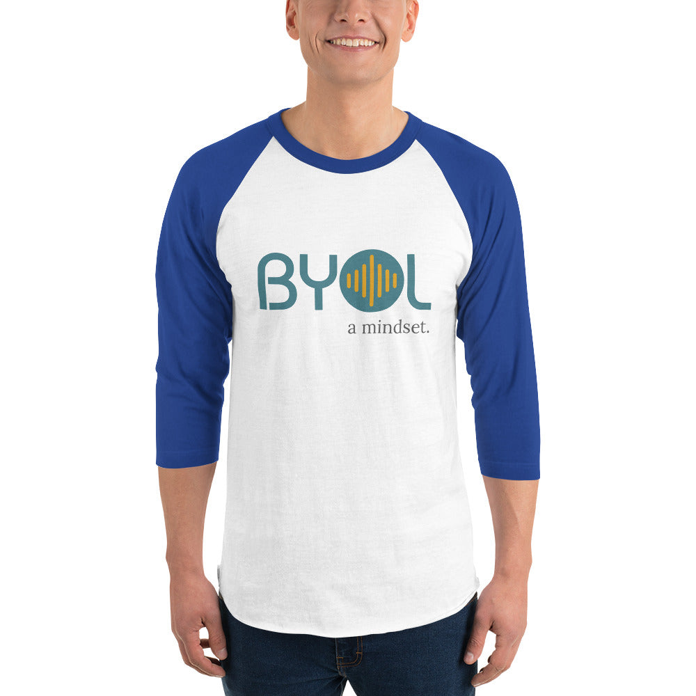 A young man wearing a white and royal blue "BYOL: a mindset" raglan T-shirt with three-quarter sleeves, smiling and standing with his arms at his sides. The T-shirt features the "BYOL" logo in teal and yellow on the front and is available in multiple colors (red, black, white, green, plus more) and sizes (S-3XL). The "be you out loud" logo is displayed at the top.