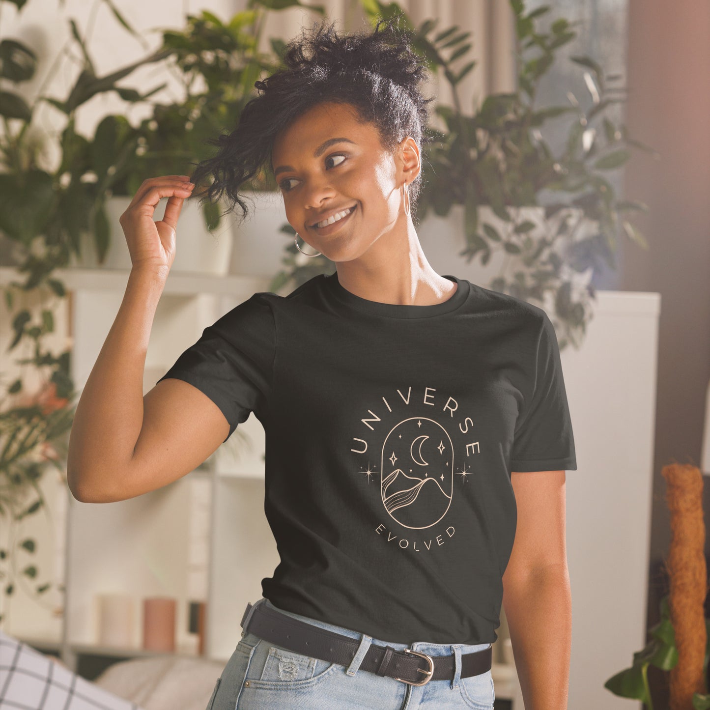 A contented woman in a black t-shirt featuring a circular graphic with the words "UNIVERSE EVOLVED" around a design of celestial bodies and a human silhouette, alluding to cosmic connection and personal growth. She's playfully touching her hair and standing in a room with a soothing ambiance created by lush indoor plants.