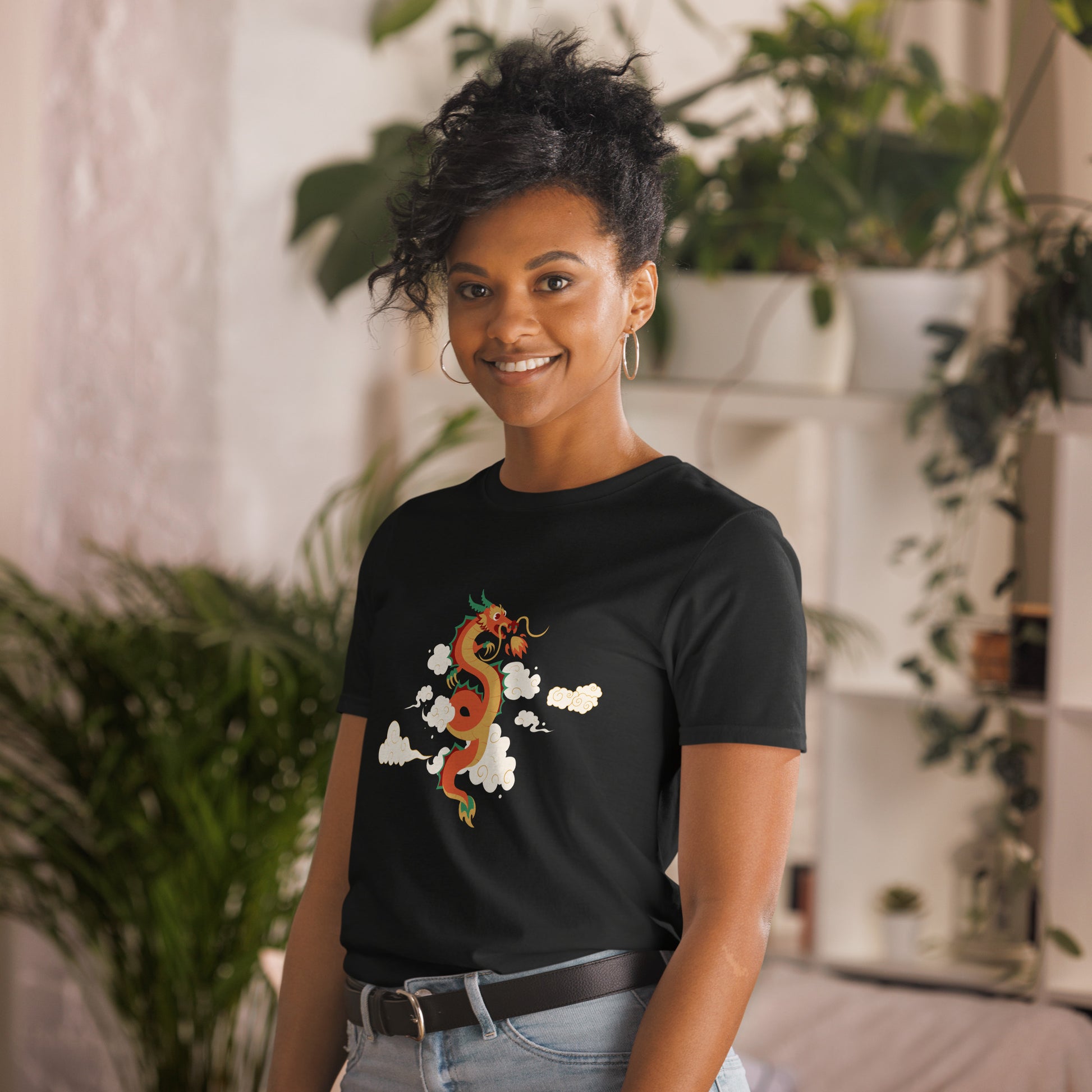 A woman with a confident smile is wearing a black t-shirt with a vibrant, multicolored dragon graphic amongst clouds, symbolizing power and mystique. She's in a cozy, plant-filled space that adds a touch of nature's tranquility to the image.