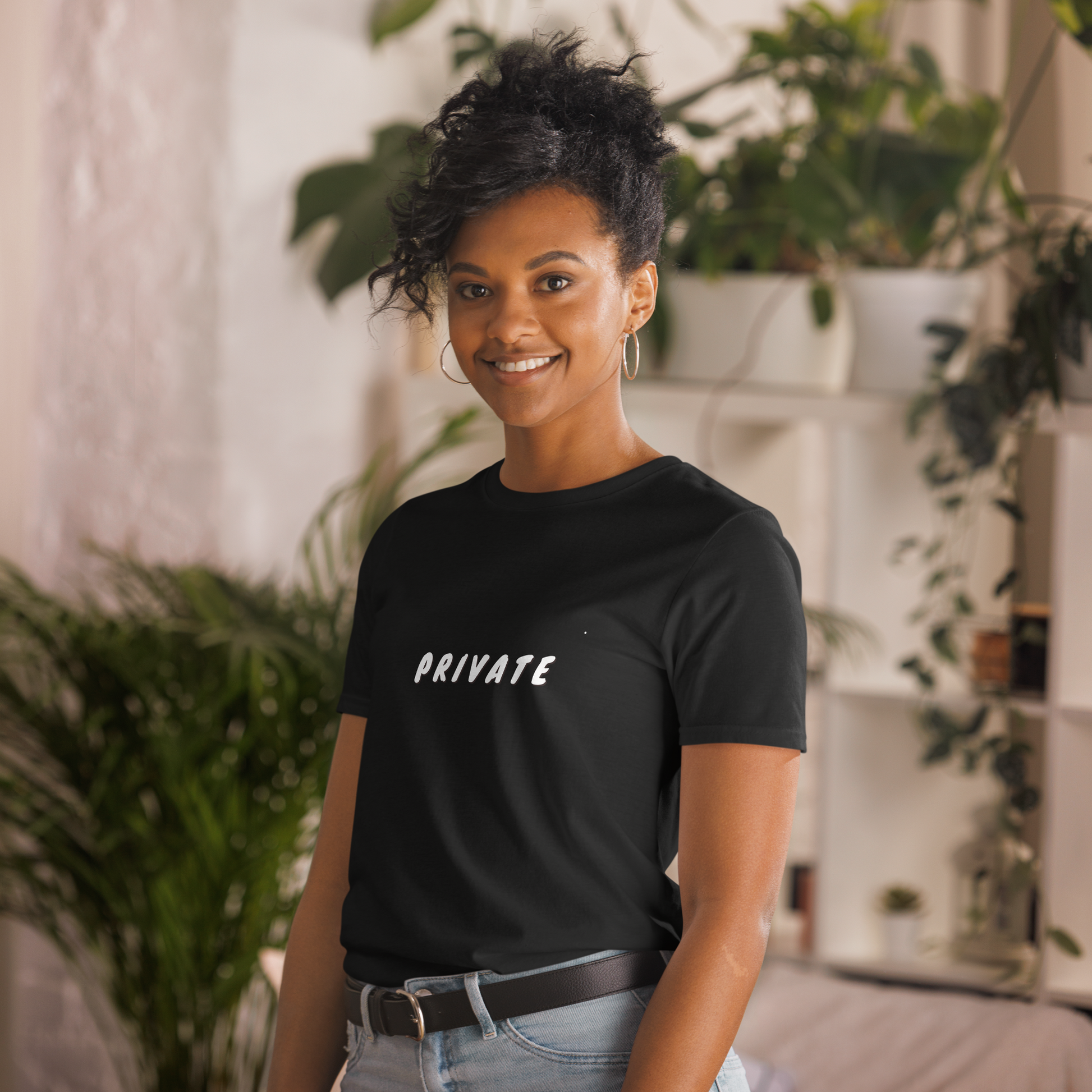 A woman with a friendly smile, wearing a black T-shirt with the word "PRIVATE" printed on it, standing in a room with houseplants.