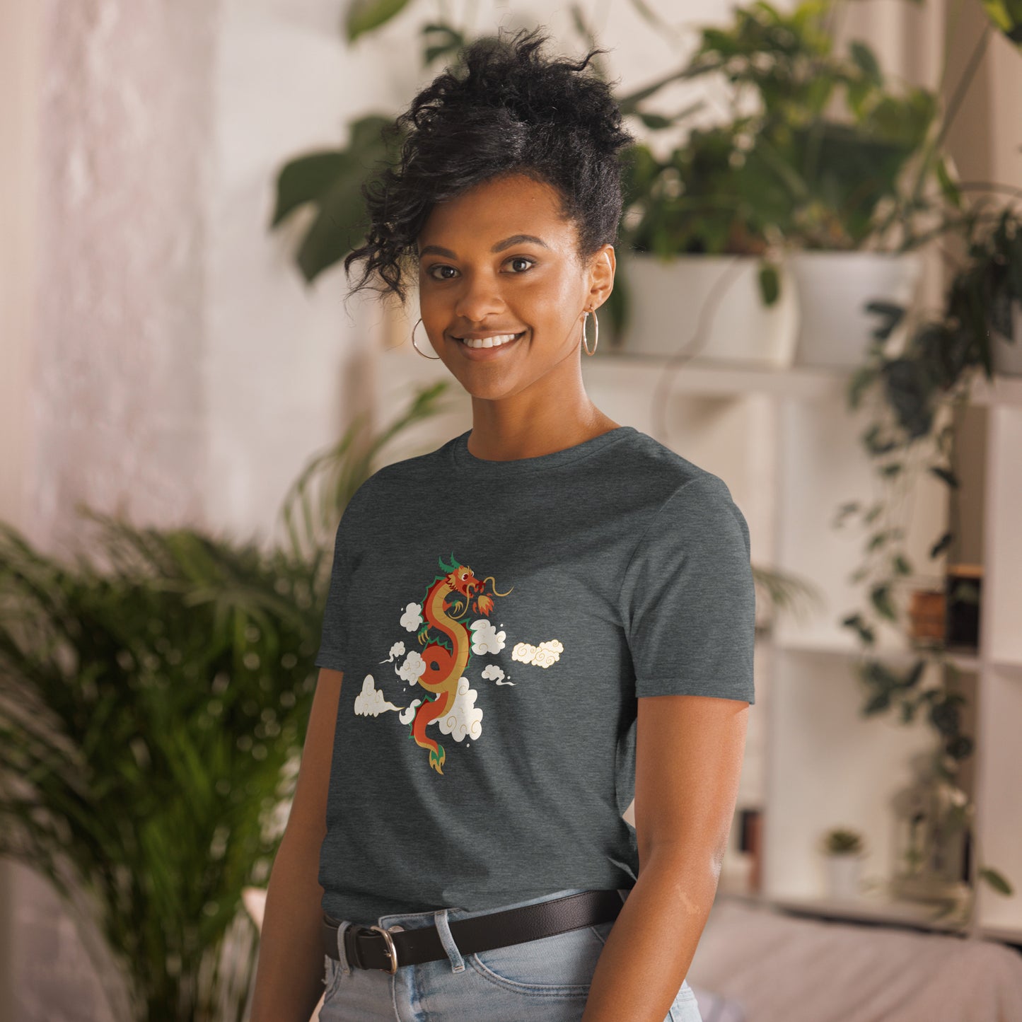 A woman with a confident smile is wearing a heathered grey t-shirt with a vibrant, multicolored dragon graphic amongst clouds, symbolizing power and mystique. She's in a cozy, plant-filled space that adds a touch of nature's tranquility to the image.