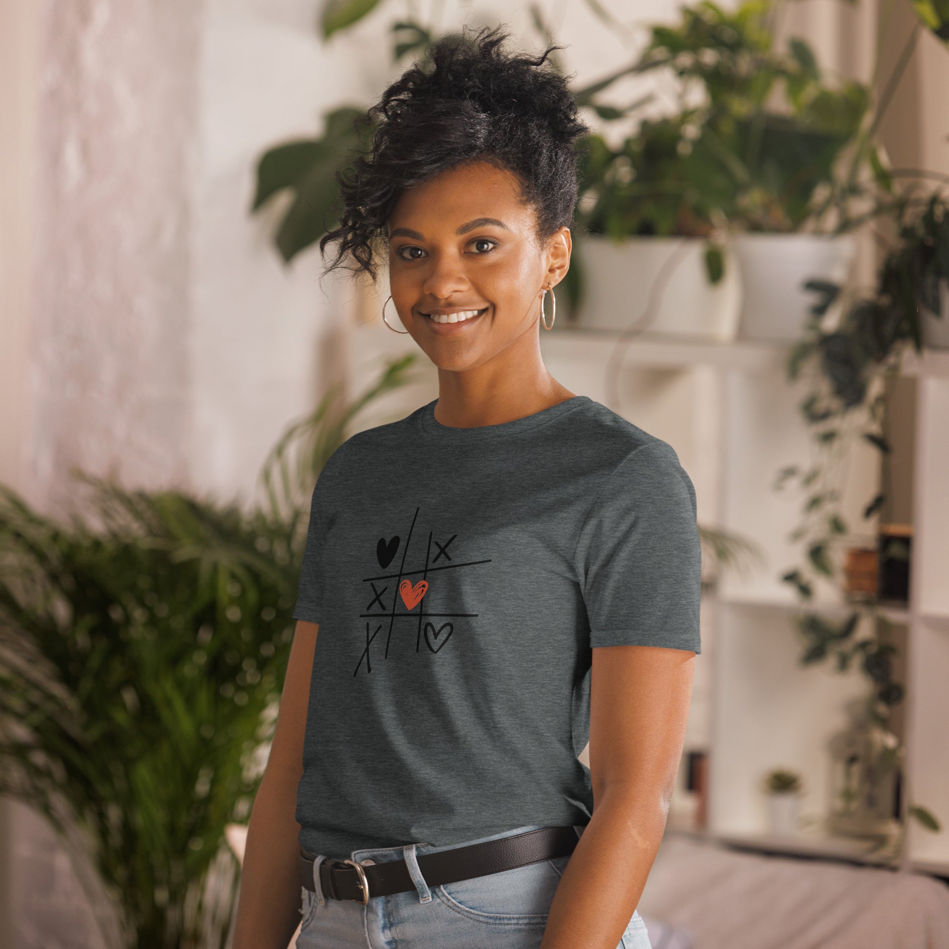 A woman with a playful hairstyle and a warm smile is wearing a dark heather grey t-shirt with a tic-tac-toe design featuring hearts and X's, creating a charming and lighthearted look. She's standing in a cozy, plant-filled room, adding to the casual and relaxed vibe.