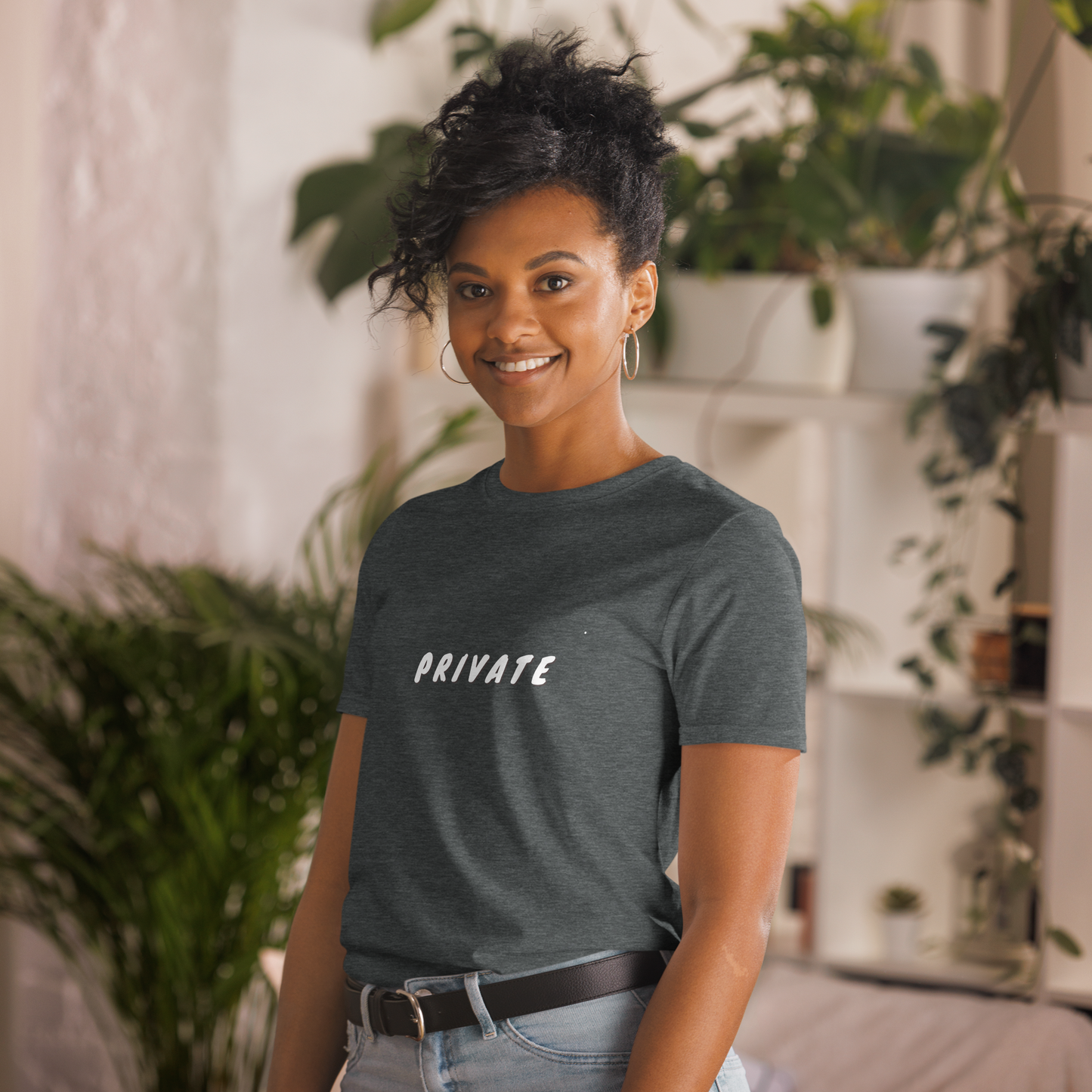 A woman with a friendly smile, wearing a heathered grey T-shirt with the word "PRIVATE" printed on it, standing in a room with houseplants.