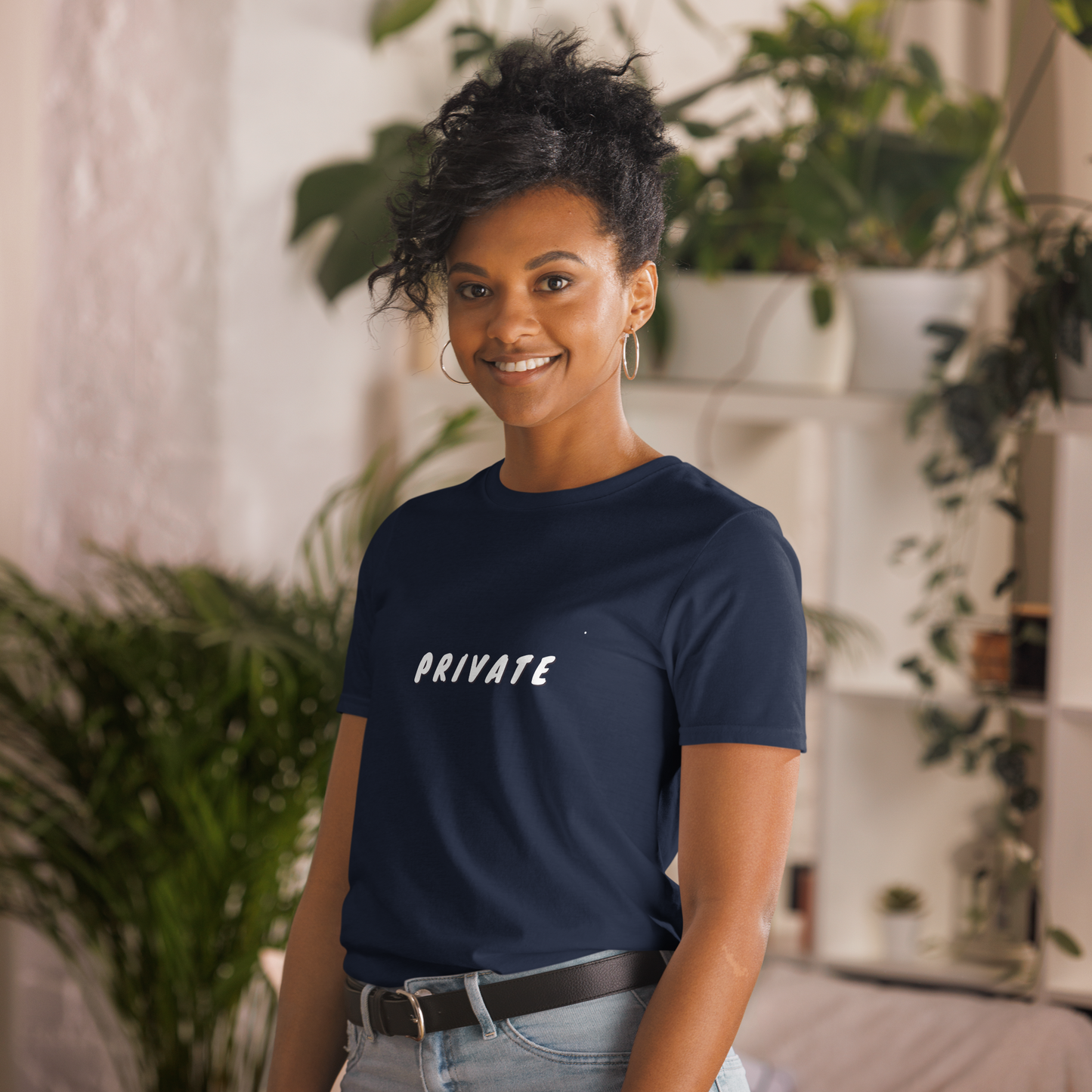 A woman with a friendly smile, wearing a navy blue T-shirt with the word "PRIVATE" printed on it, standing in a room with houseplants.