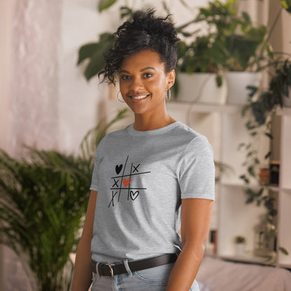 A woman with a playful hairstyle and a warm smile is wearing a heather grey t-shirt with a tic-tac-toe design featuring hearts and X's, creating a charming and lighthearted look. She's standing in a cozy, plant-filled room, adding to the casual and relaxed vibe.