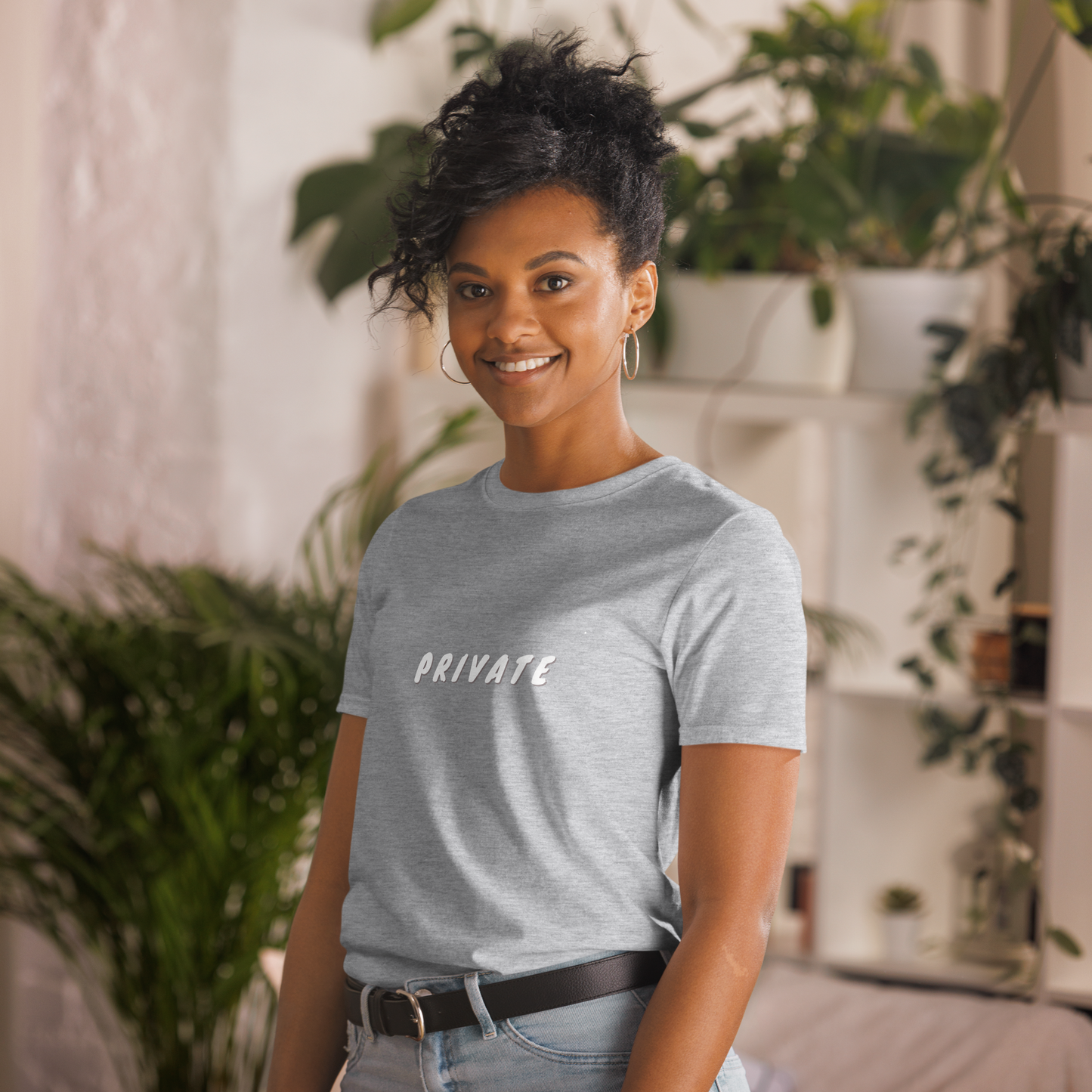 A woman with a friendly smile, wearing a light heathered grey T-shirt with the word "PRIVATE" printed on it, standing in a room with houseplants.