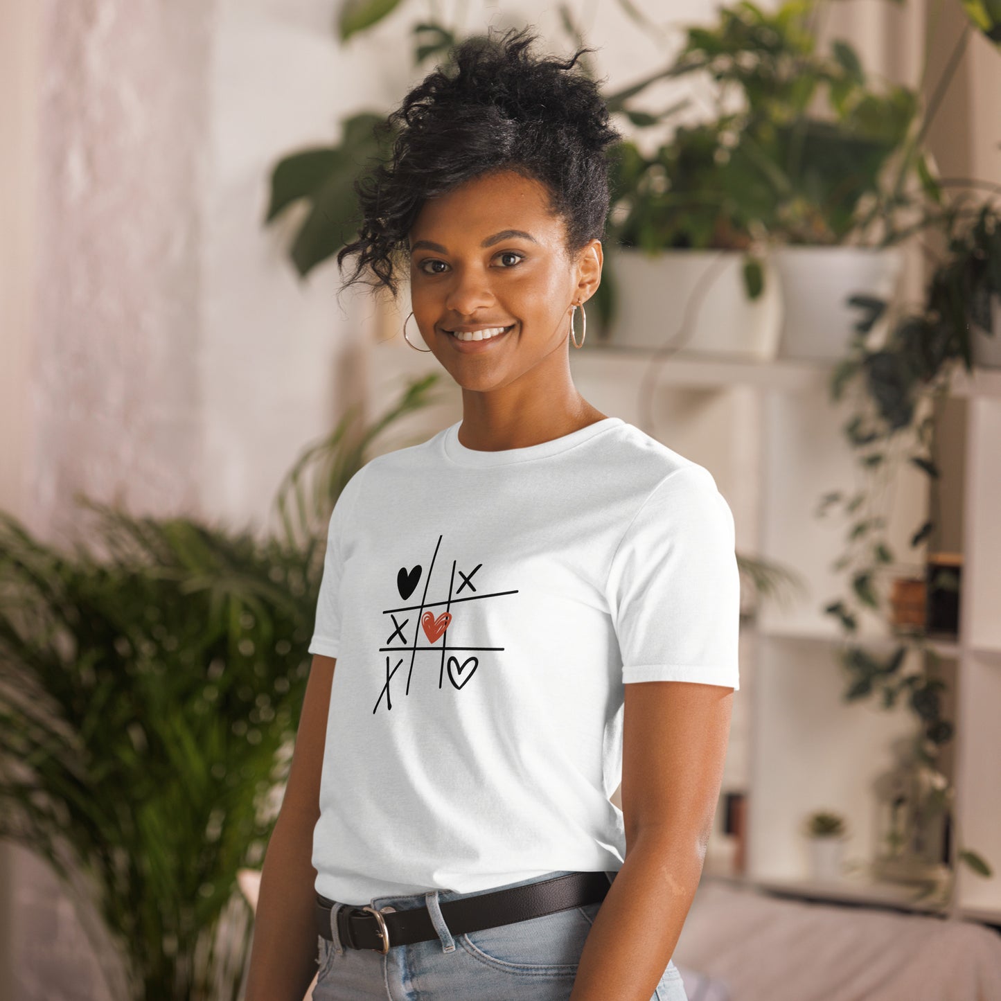 A woman with a playful hairstyle and a warm smile is wearing a white t-shirt with a tic-tac-toe design featuring hearts and X's, creating a charming and lighthearted look. She's standing in a cozy, plant-filled room, adding to the casual and relaxed vibe.