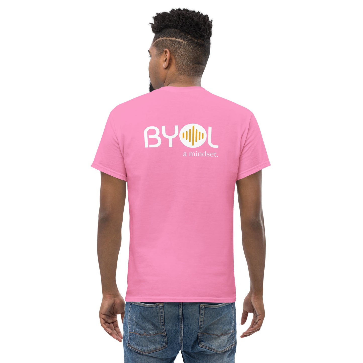 A young man with short curly hair wearing an pink "BYOL: a mindset" T-shirt, viewed from the back. The T-shirt features the "BYOL" logo in white and yellow on the back and is available in multiple colors (maroon, black, brown, gold, plus more) and sizes (S-3XL). The "be you out loud" logo is displayed at the top.