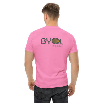 A young man with short blonde hair wearing a pink "BYOL: a mindset" T-shirt, viewed from the back. The T-shirt features the "BYOL" logo in gray and yellow on the back and is available in multiple colors (maroon, black, brown, gold, plus more) and sizes (S-3XL). The "be you out loud" logo is displayed at the top.