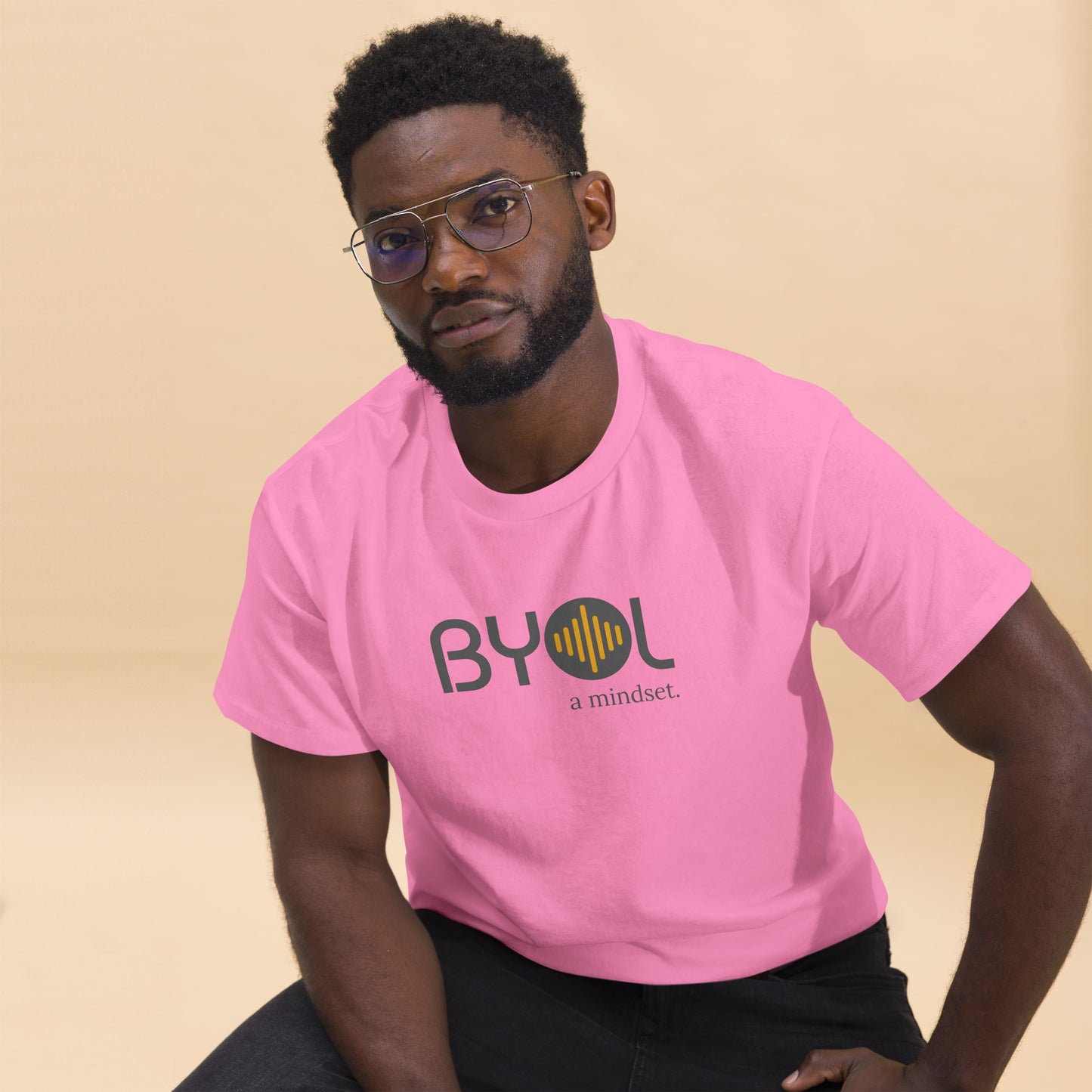 A young man with short curly hair and glasses wearing a pink "BYOL: a mindset" T-shirt, sitting down. The T-shirt features the "BYOL" logo in gray and yellow on the front and is available in multiple colors (maroon, black, brown, gold, plus more) and sizes (S-3XL). The "be you out loud" logo is displayed at the top.