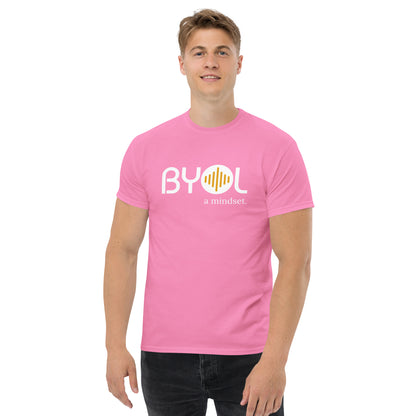 A young man with short blonde hair wearing an pink "BYOL: a mindset" T-shirt, smiling and standing with his arms at his sides. The T-shirt features the "BYOL" logo in white and yellow on the front and is available in multiple colors (maroon, black, brown, gold, plus more) and sizes (S-3XL). The "be you out loud" logo is displayed at the top.