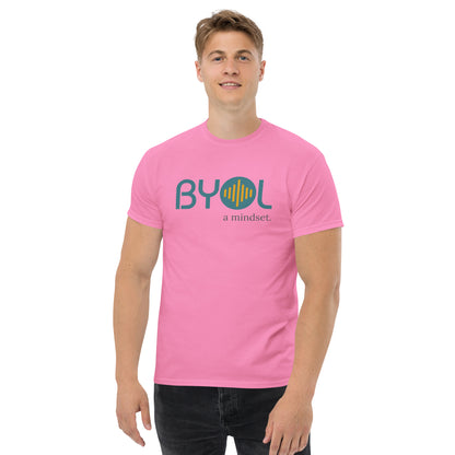 A young man with short blonde hair wearing a pink "BYOL: a mindset" T-shirt, smiling and standing with his arms at his sides. The T-shirt features the "BYOL" logo in teal and yellow on the front and is available in multiple colors (maroon, black, brown, gold, plus more) and sizes (S-3XL). The "be you out loud" logo is displayed at the top.