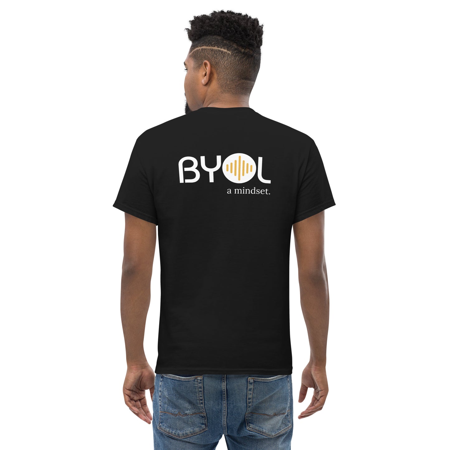 A young man with short curly hair wearing an black "BYOL: a mindset" T-shirt, viewed from the back. The T-shirt features the "BYOL" logo in white and yellow on the back and is available in multiple colors (maroon, black, brown, gold, plus more) and sizes (S-3XL). The "be you out loud" logo is displayed at the top.