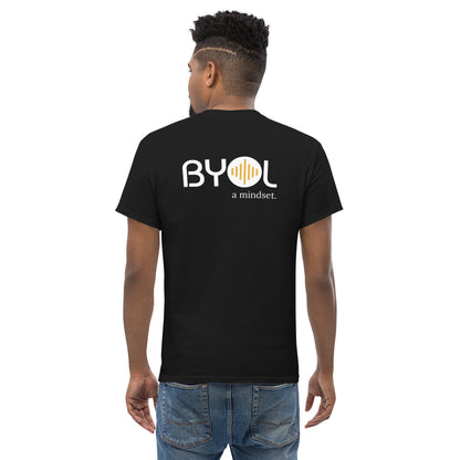 A young man with short curly hair wearing an black "BYOL: a mindset" T-shirt, viewed from the back. The T-shirt features the "BYOL" logo in white and yellow on the back and is available in multiple colors (maroon, black, brown, gold, plus more) and sizes (S-3XL). The "be you out loud" logo is displayed at the top.