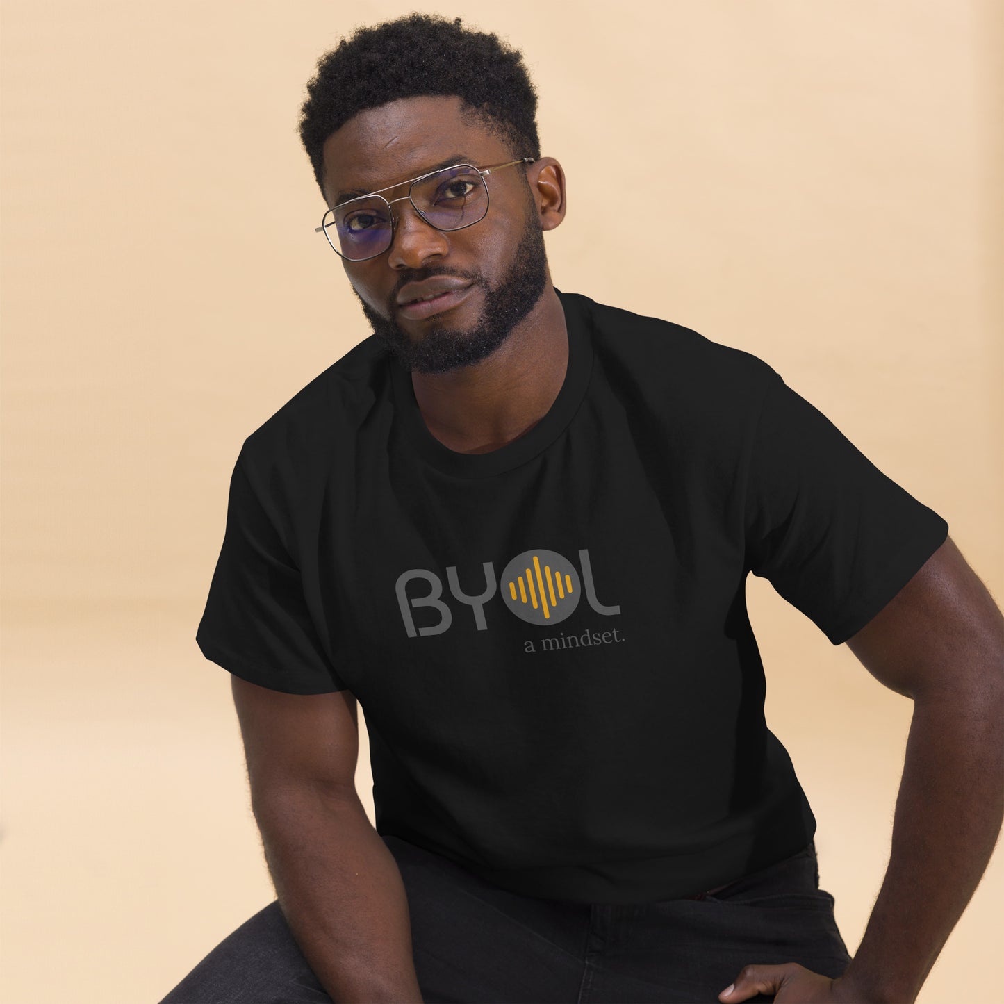 A young man with short curly hair and glasses wearing a black "BYOL: a mindset" T-shirt, sitting down. The T-shirt features the "BYOL" logo in gray and yellow on the front and is available in multiple colors (maroon, black, brown, gold, plus more) and sizes (S-3XL). The "be you out loud" logo is displayed at the top.