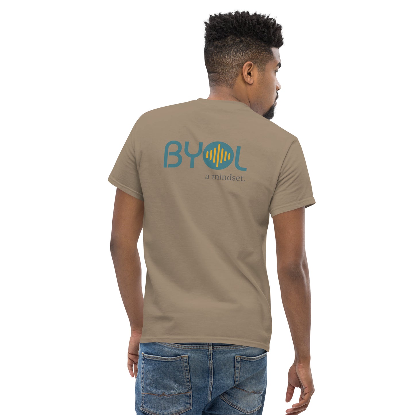 A young man with short hair wearing a savana brown "BYOL: a mindset" T-shirt, viewed from the back. The T-shirt features the "BYOL" logo in teal and yellow on the back and is available in multiple colors (maroon, black, brown, gold, plus more) and sizes (S-3XL). The "be you out loud" logo is displayed at the top.