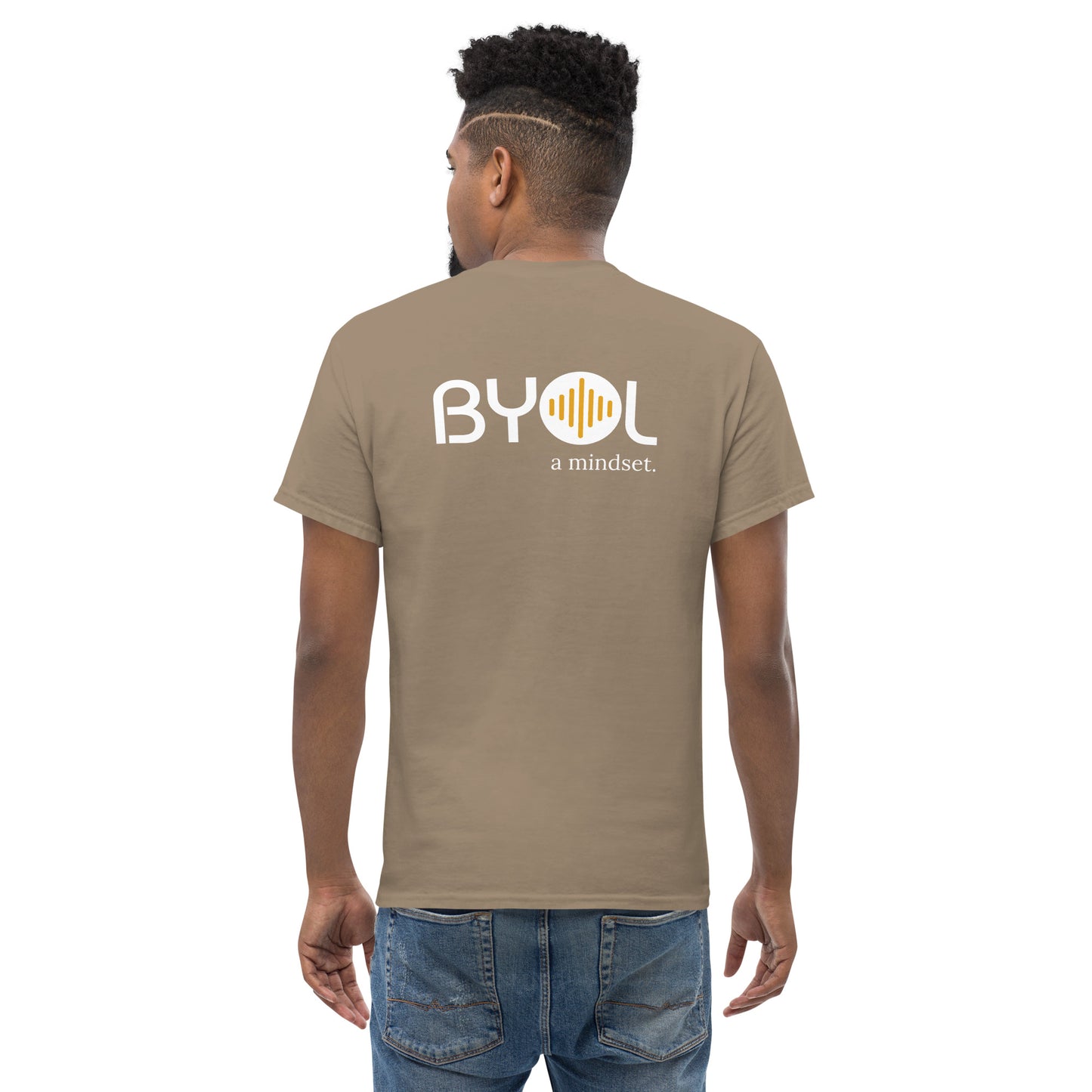 A young man with short curly hair wearing an savana brown "BYOL: a mindset" T-shirt, viewed from the back. The T-shirt features the "BYOL" logo in white and yellow on the back and is available in multiple colors (maroon, black, brown, gold, plus more) and sizes (S-3XL). The "be you out loud" logo is displayed at the top.