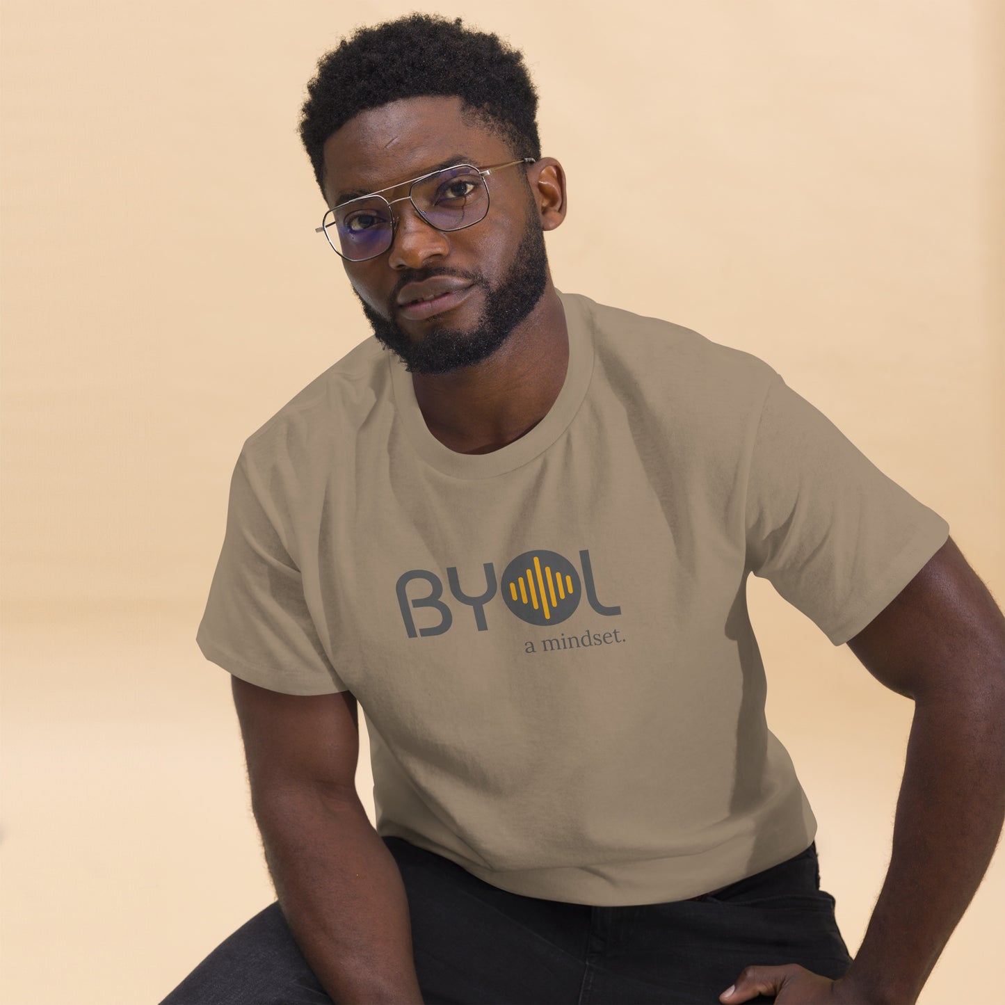 A young man with short curly hair and glasses wearing a brown savana "BYOL: a mindset" T-shirt, sitting down. The T-shirt features the "BYOL" logo in gray and yellow on the front and is available in multiple colors (maroon, black, brown, gold, plus more) and sizes (S-3XL). The "be you out loud" logo is displayed at the top.