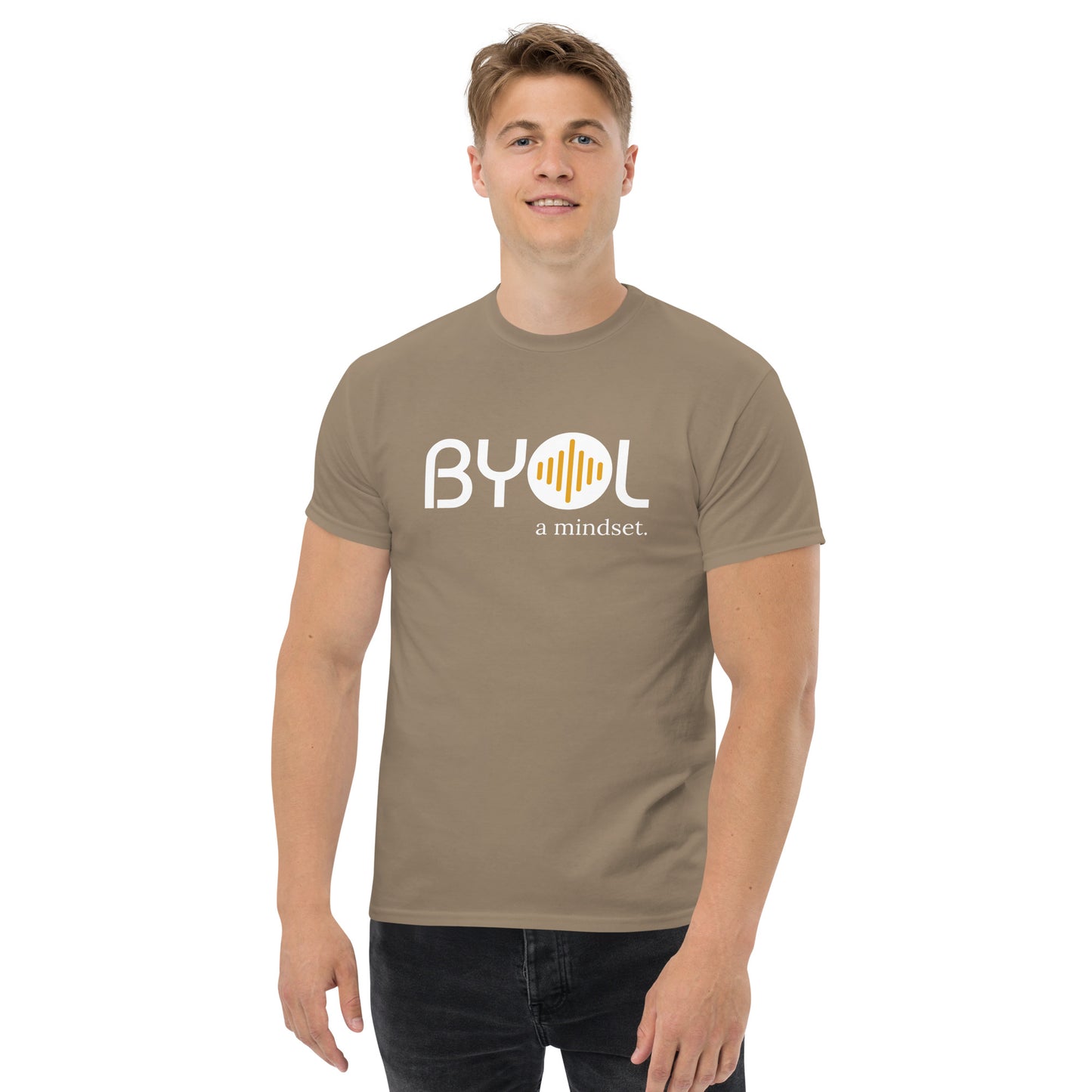 A young man with short blonde hair wearing an savana brown "BYOL: a mindset" T-shirt, smiling and standing with his arms at his sides. The T-shirt features the "BYOL" logo in white and yellow on the front and is available in multiple colors (maroon, black, brown, gold, plus more) and sizes (S-3XL). The "be you out loud" logo is displayed at the top.