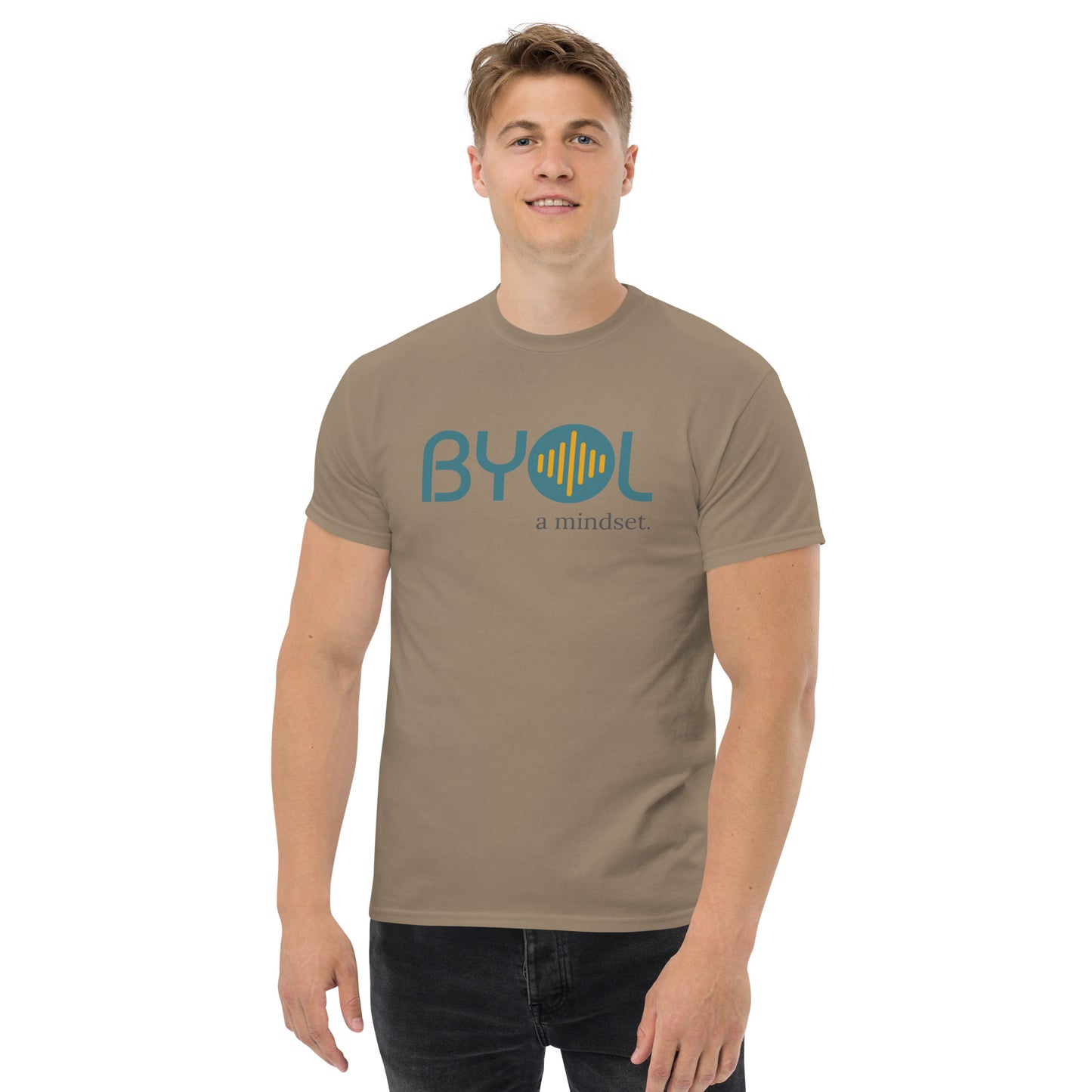 A young man with short blonde hair wearing a savana brown "BYOL: a mindset" T-shirt, smiling and standing with his arms at his sides. The T-shirt features the "BYOL" logo in teal and yellow on the front and is available in multiple colors (maroon, black, brown, gold, plus more) and sizes (S-3XL). The "be you out loud" logo is displayed at the top.