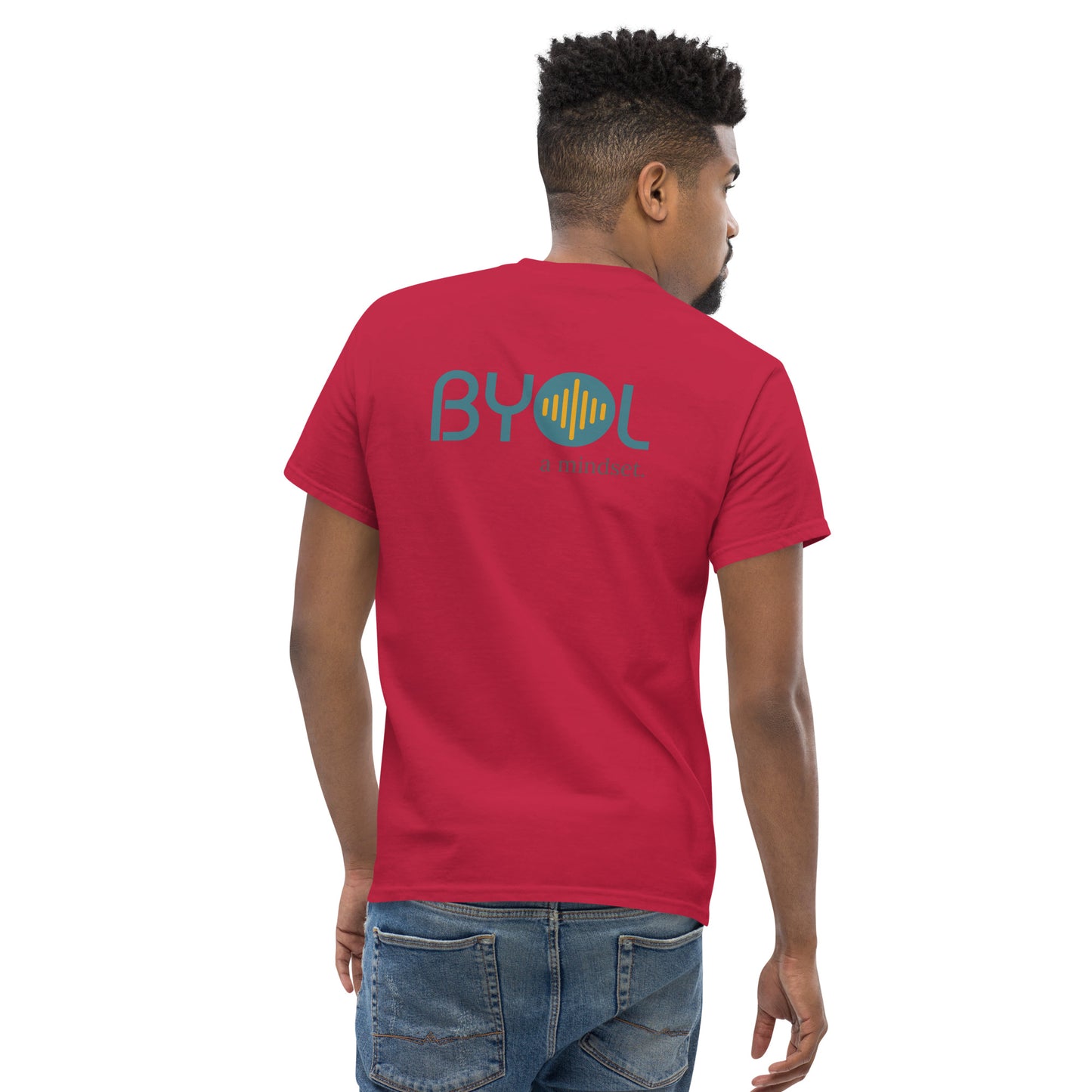 A young man with short hair wearing a cardinal red "BYOL: a mindset" T-shirt, viewed from the back. The T-shirt features the "BYOL" logo in teal and yellow on the back and is available in multiple colors (maroon, black, brown, gold, plus more) and sizes (S-3XL). The "be you out loud" logo is displayed at the top.