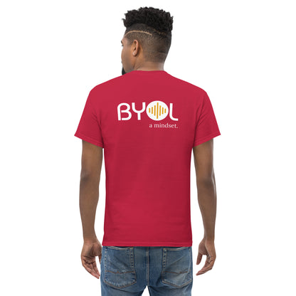 A young man with short curly hair wearing an cardinal red "BYOL: a mindset" T-shirt, viewed from the back. The T-shirt features the "BYOL" logo in white and yellow on the back and is available in multiple colors (maroon, black, brown, gold, plus more) and sizes (S-3XL). The "be you out loud" logo is displayed at the top.