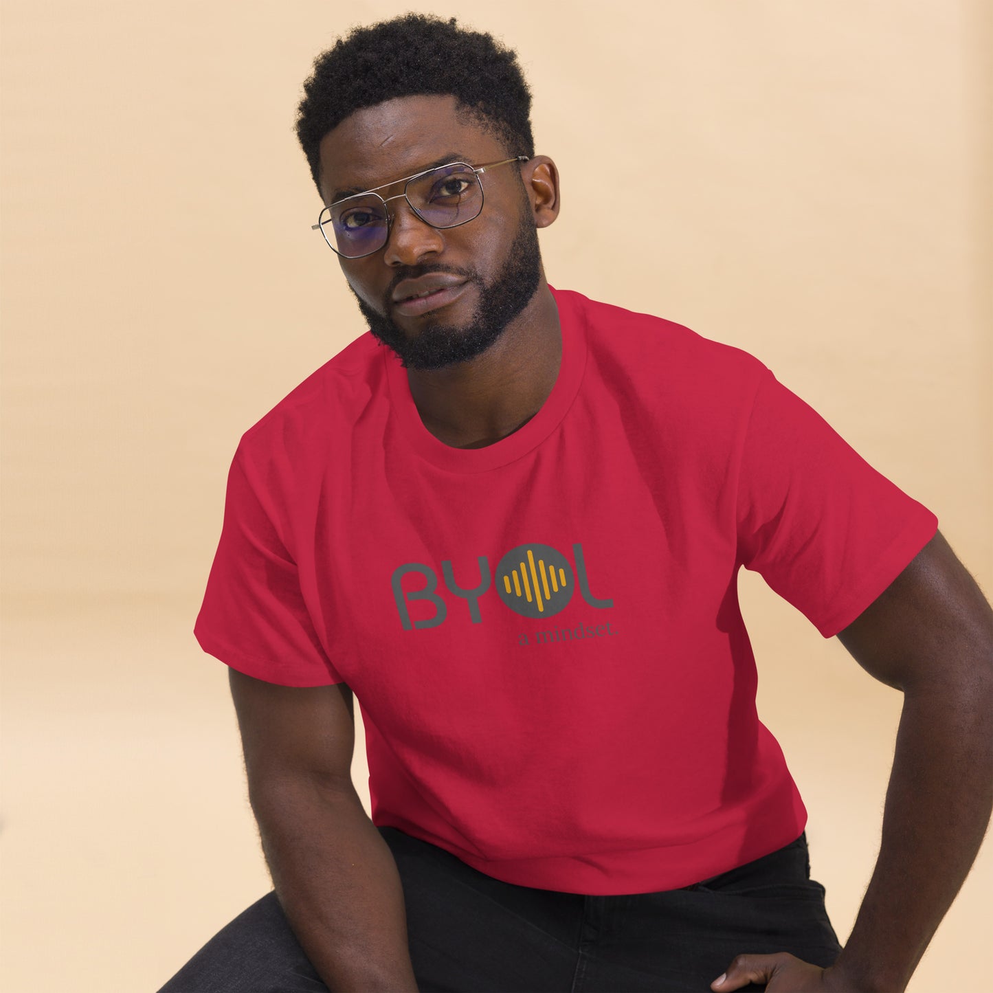 A young man with short curly hair and glasses wearing a cardinal red "BYOL: a mindset" T-shirt, sitting down. The T-shirt features the "BYOL" logo in gray and yellow on the front and is available in multiple colors (maroon, black, brown, gold, plus more) and sizes (S-3XL). The "be you out loud" logo is displayed at the top.