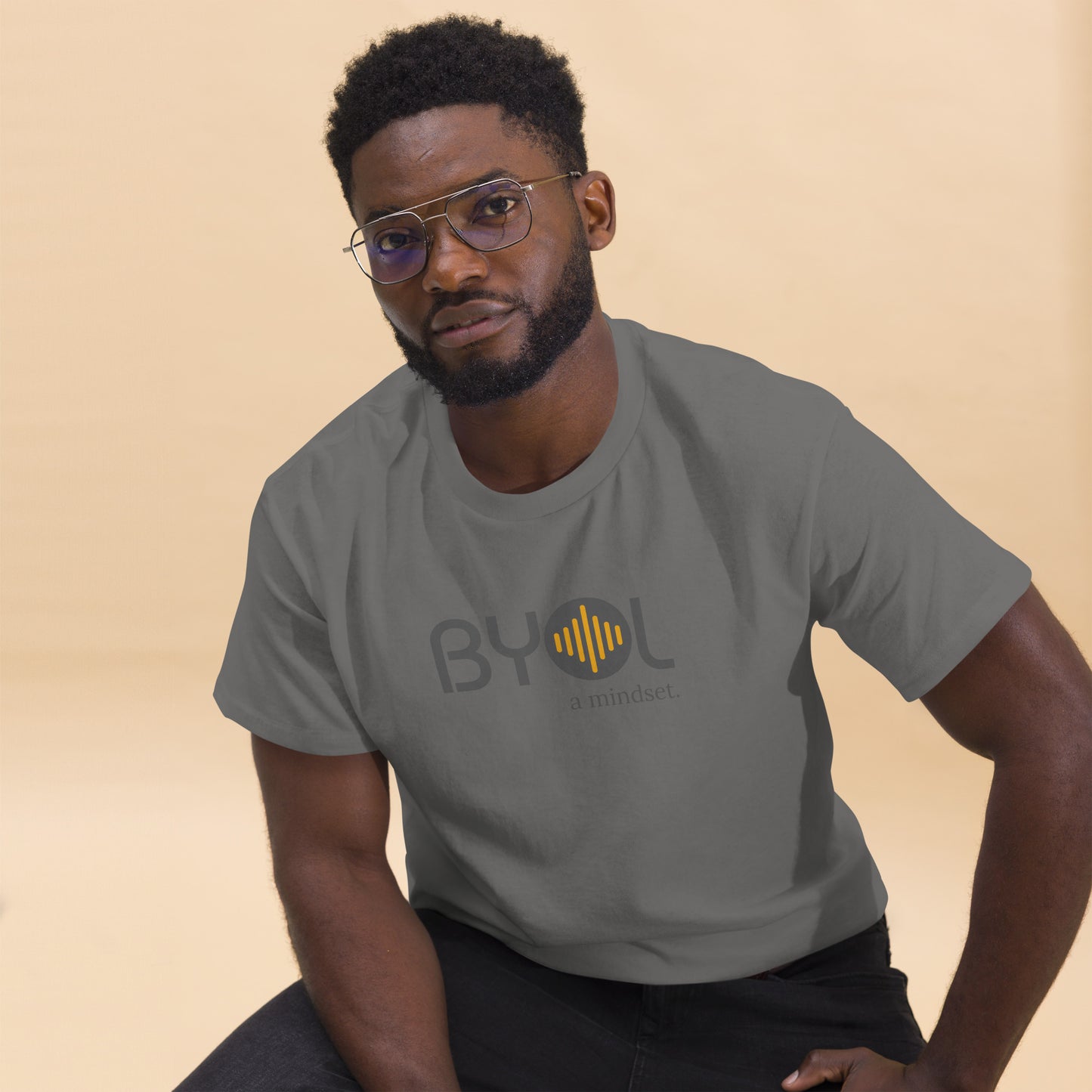 A young man with short curly hair and glasses wearing a charcoal "BYOL: a mindset" T-shirt, sitting down. The T-shirt features the "BYOL" logo in gray and yellow on the front and is available in multiple colors (maroon, black, brown, gold, plus more) and sizes (S-3XL). The "be you out loud" logo is displayed at the top.