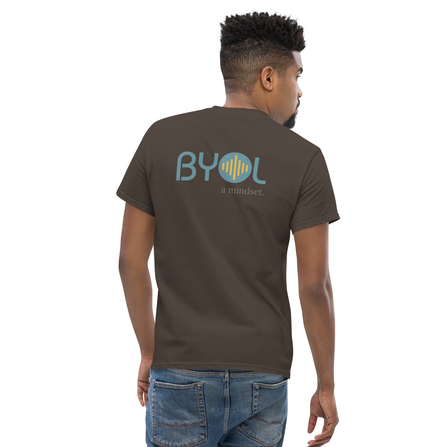 A young man with short hair wearing a dark brown "BYOL: a mindset" T-shirt, viewed from the back. The T-shirt features the "BYOL" logo in teal and yellow on the back and is available in multiple colors (maroon, black, brown, gold, plus more) and sizes (S-3XL). The "be you out loud" logo is displayed at the top.