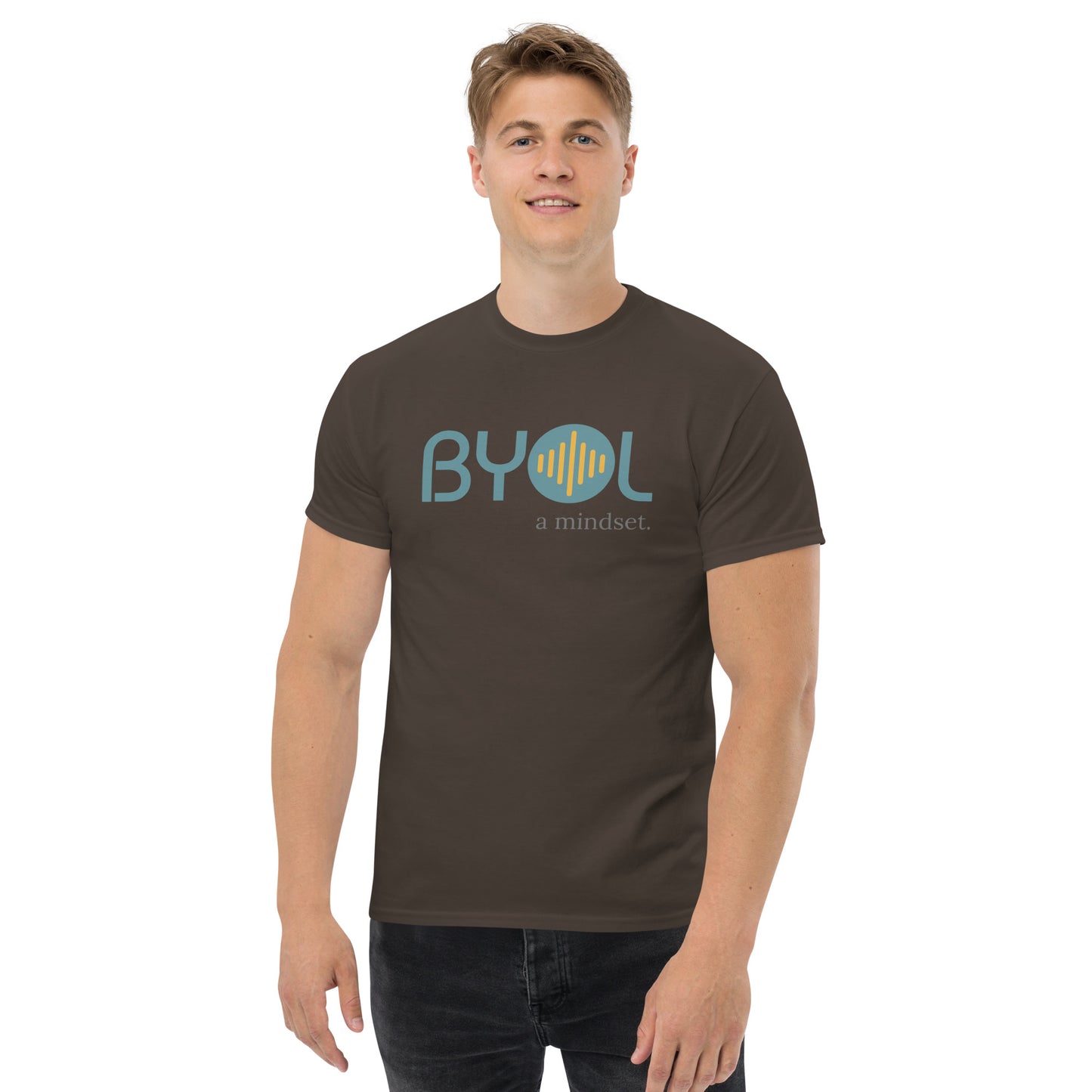 A young man with short blonde hair wearing a dark chocolate "BYOL: a mindset" T-shirt, smiling and standing with his arms at his sides. The T-shirt features the "BYOL" logo in teal and yellow on the front and is available in multiple colors (maroon, black, brown, gold, plus more) and sizes (S-3XL). The "be you out loud" logo is displayed at the top.