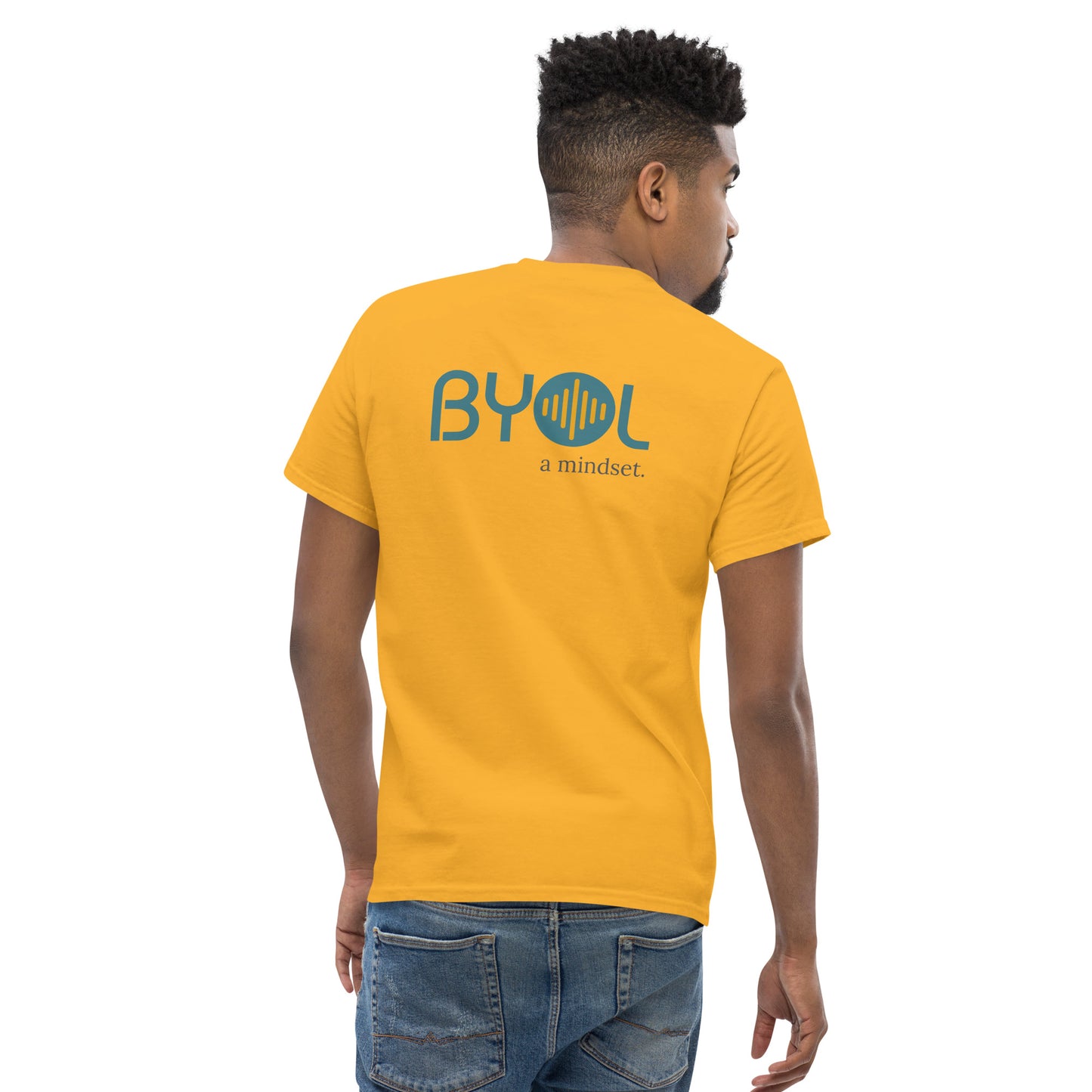 A young man with short hair wearing a yellow "BYOL: a mindset" T-shirt, viewed from the back. The T-shirt features the "BYOL" logo in teal and yellow on the back and is available in multiple colors (maroon, black, brown, gold, plus more) and sizes (S-3XL). The "be you out loud" logo is displayed at the top.