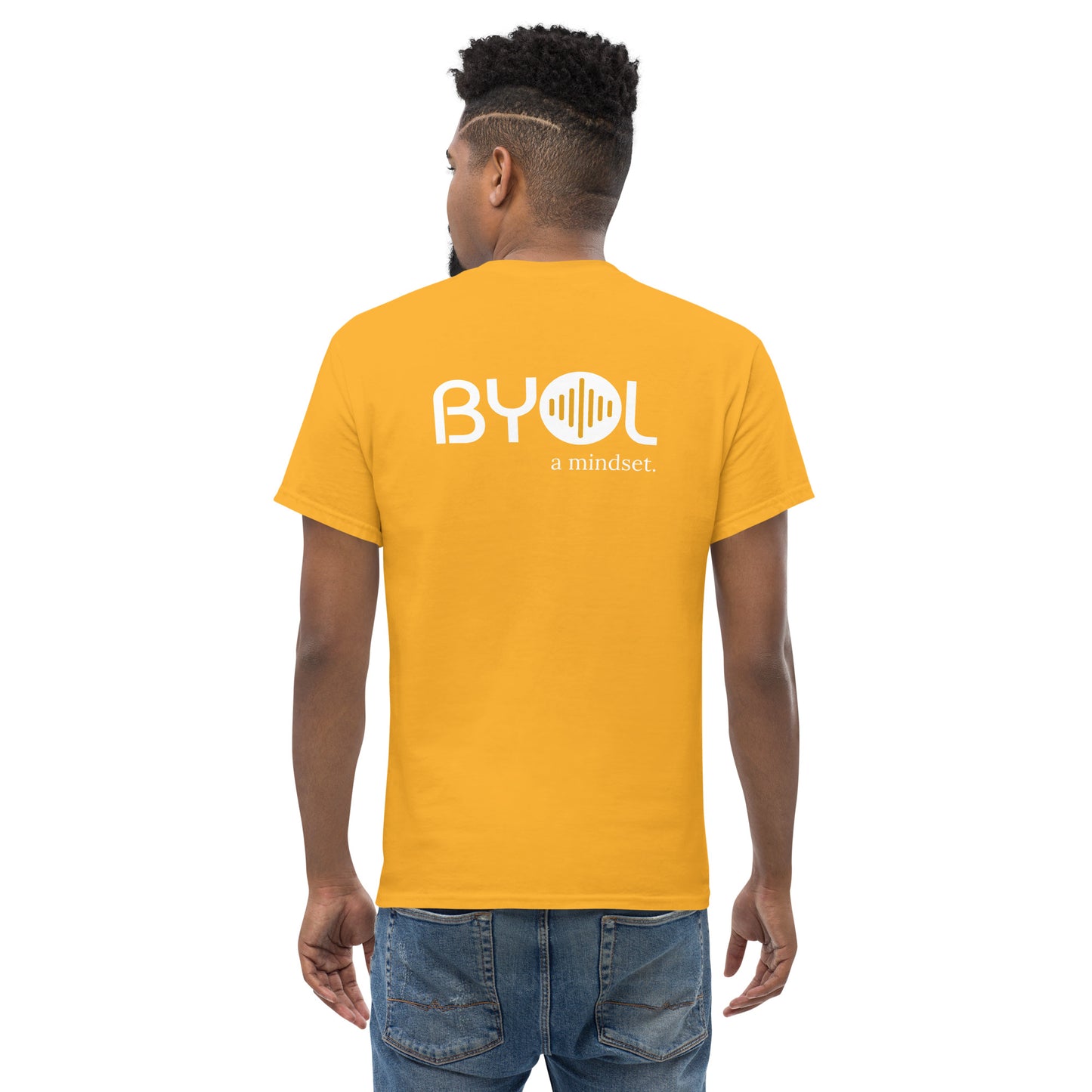 A young man with short curly hair wearing an yellow "BYOL: a mindset" T-shirt, viewed from the back. The T-shirt features the "BYOL" logo in white and yellow on the back and is available in multiple colors (maroon, black, brown, gold, plus more) and sizes (S-3XL). The "be you out loud" logo is displayed at the top.