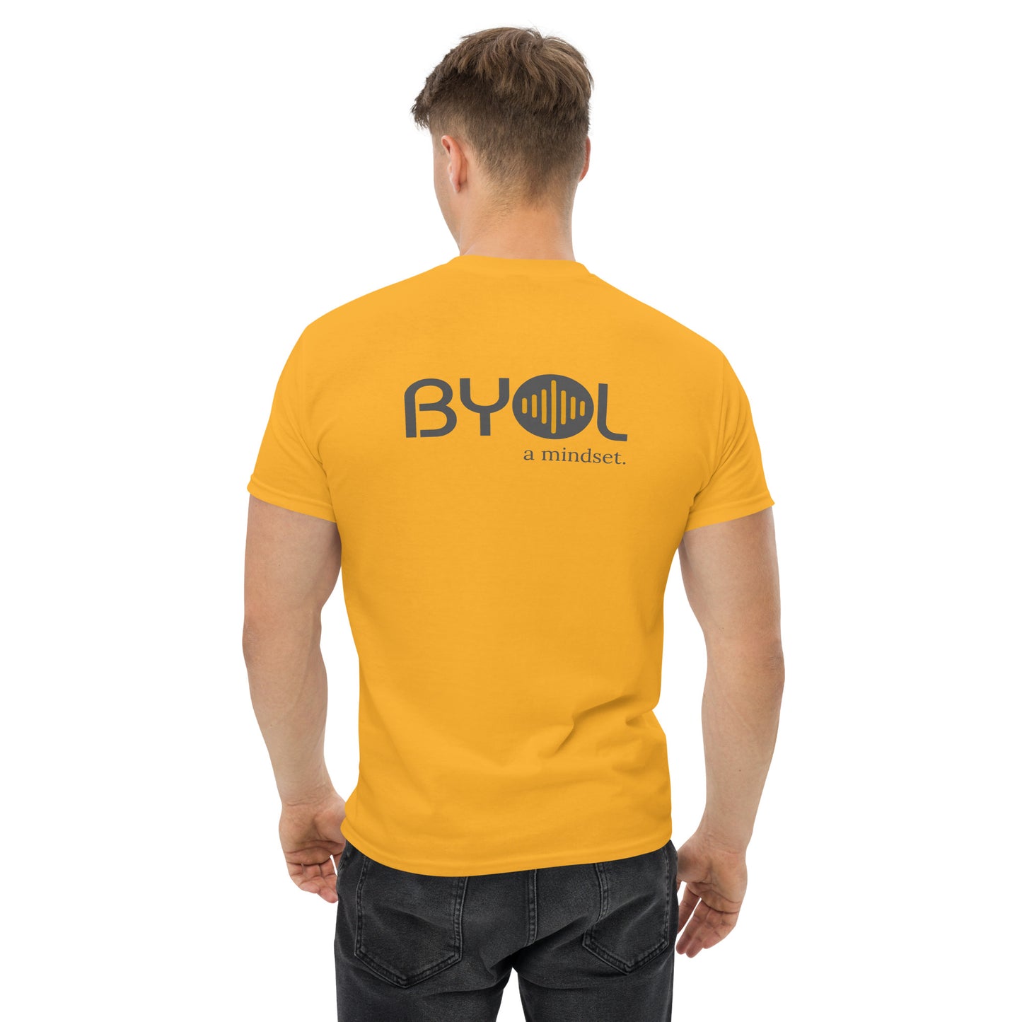 A young man with short blonde hair wearing a yellow "BYOL: a mindset" T-shirt, viewed from the back. The T-shirt features the "BYOL" logo in gray and yellow on the back and is available in multiple colors (maroon, black, brown, gold, plus more) and sizes (S-3XL). The "be you out loud" logo is displayed at the top.