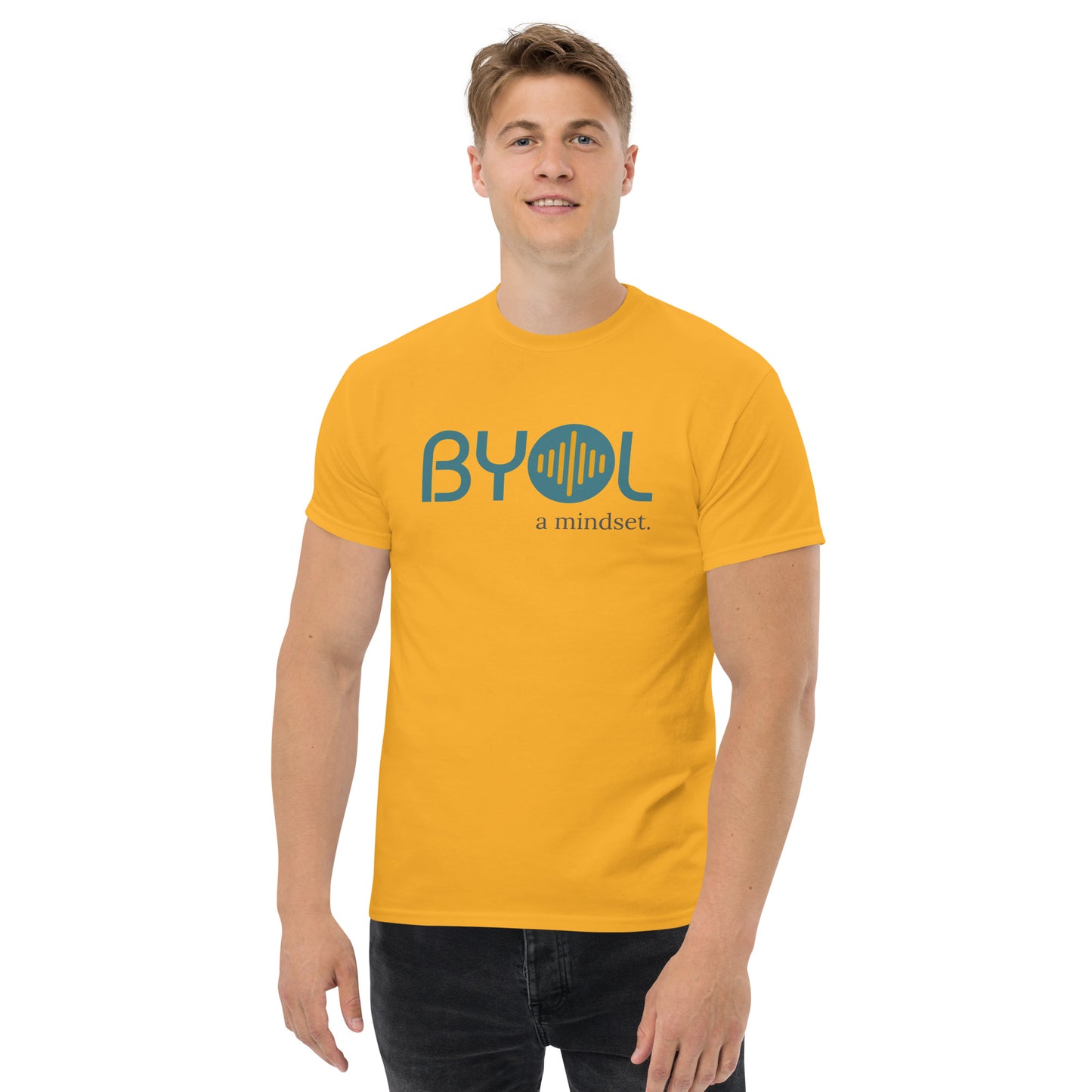 A young man with short blonde hair wearing a yellow "BYOL: a mindset" T-shirt, smiling and standing with his arms at his sides. The T-shirt features the "BYOL" logo in teal and yellow on the front and is available in multiple colors (maroon, black, brown, gold, plus more) and sizes (S-3XL). The "be you out loud" logo is displayed at the top.