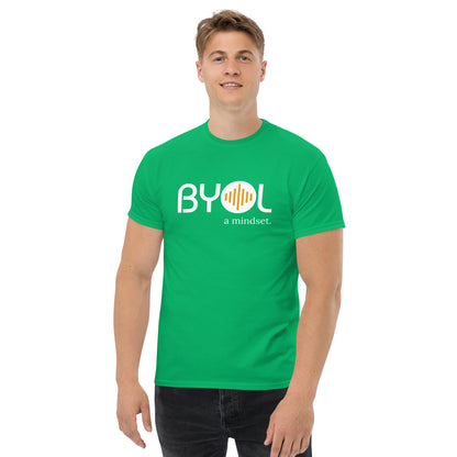 A young man with short blonde hair wearing an green "BYOL: a mindset" T-shirt, smiling and standing with his arms at his sides. The T-shirt features the "BYOL" logo in white and yellow on the front and is available in multiple colors (maroon, black, brown, gold, plus more) and sizes (S-3XL). The "be you out loud" logo is displayed at the top.